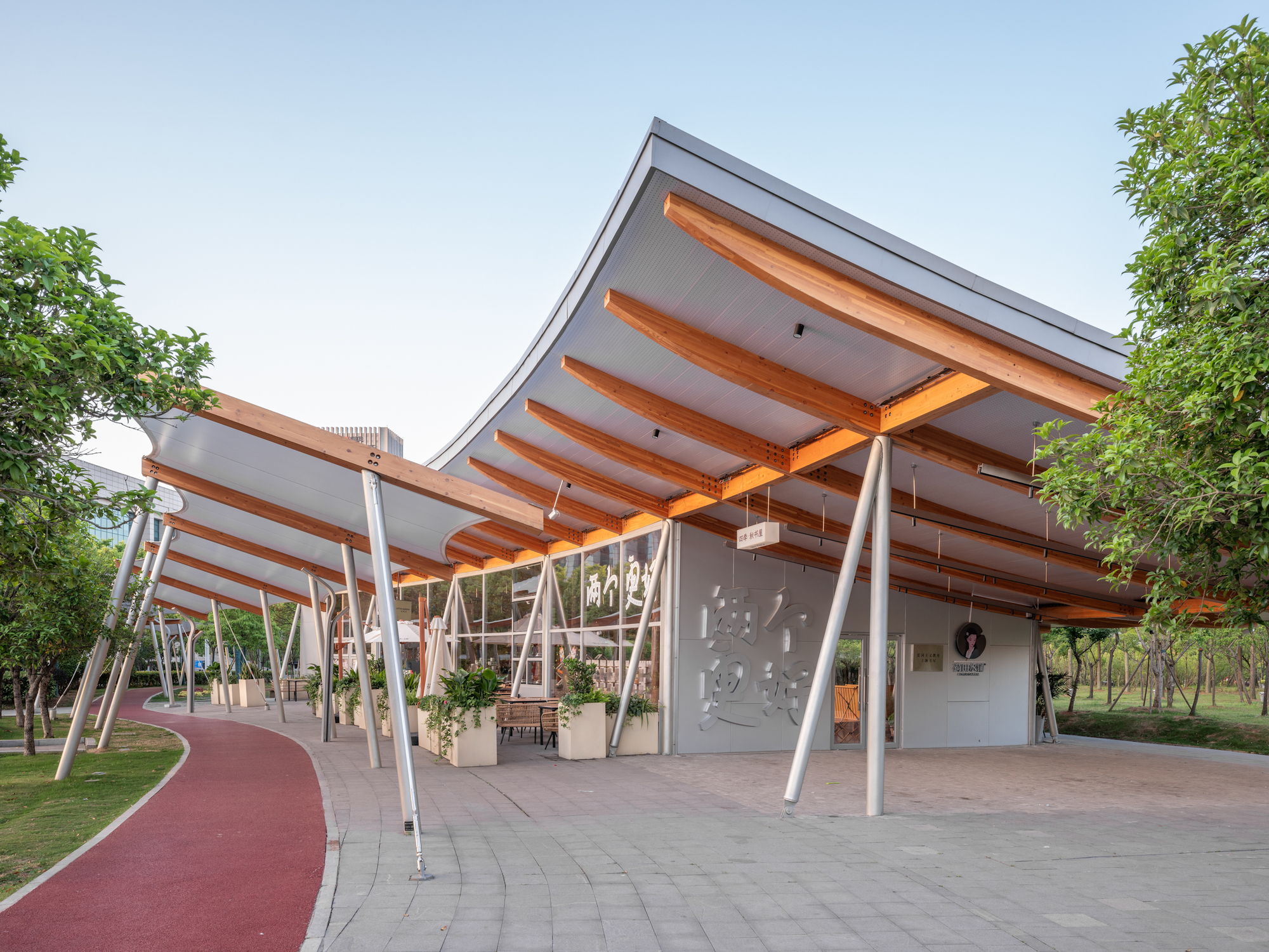 Curved canopy roof with exposed timber rafters on slender steel columns beside a red running track in late afternoon light