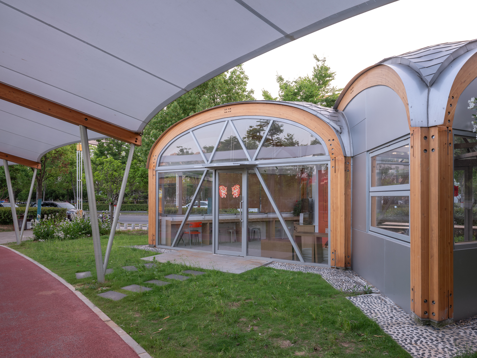 Arched timber-framed glass pavilion with diagonal bracing, seen from under the canopy across a lawn with stepping stones and pebble edging