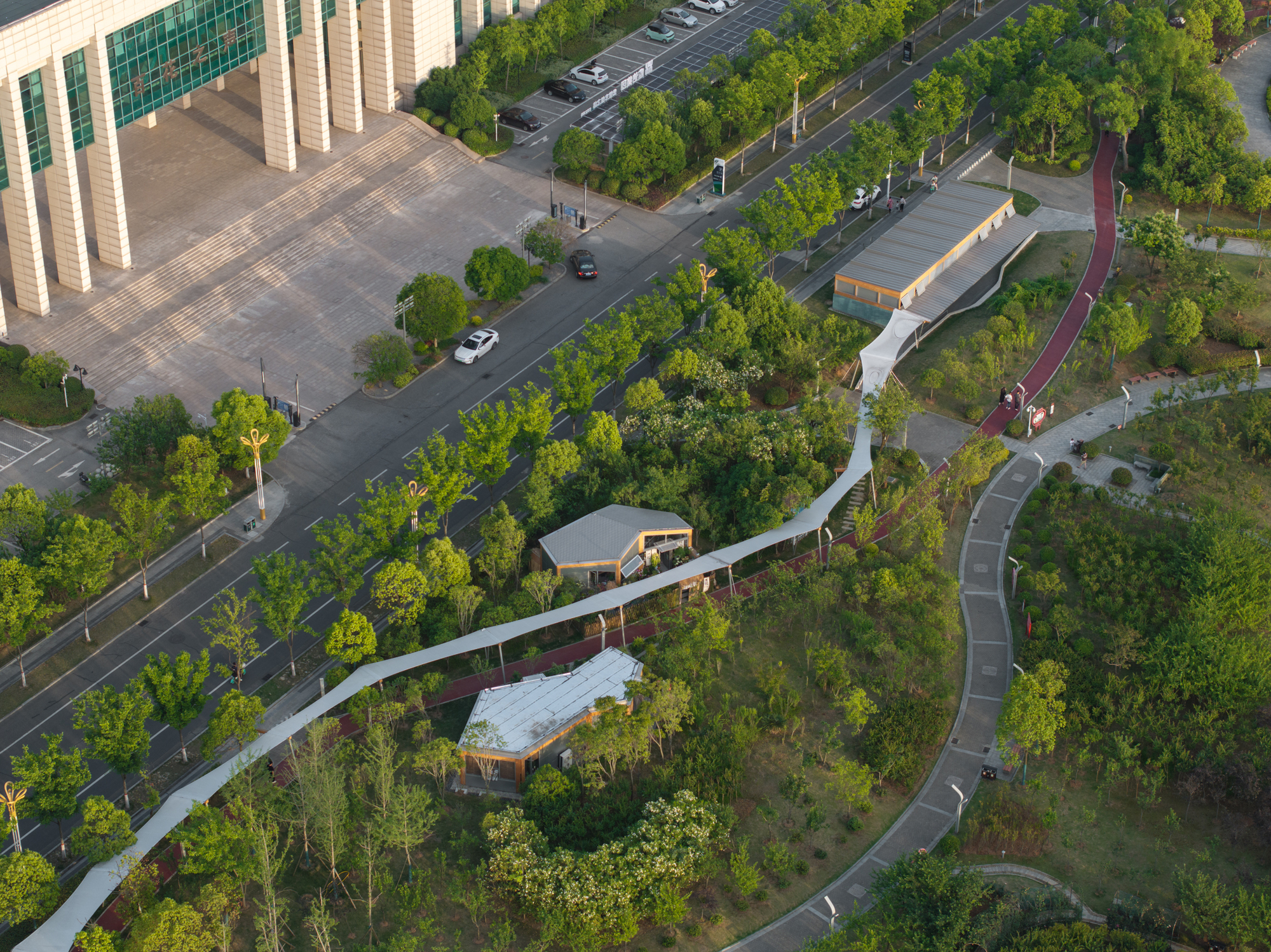 Aerial view of a ribbon-like white canopy path winding between small pavilions and trees beside a road at dusk