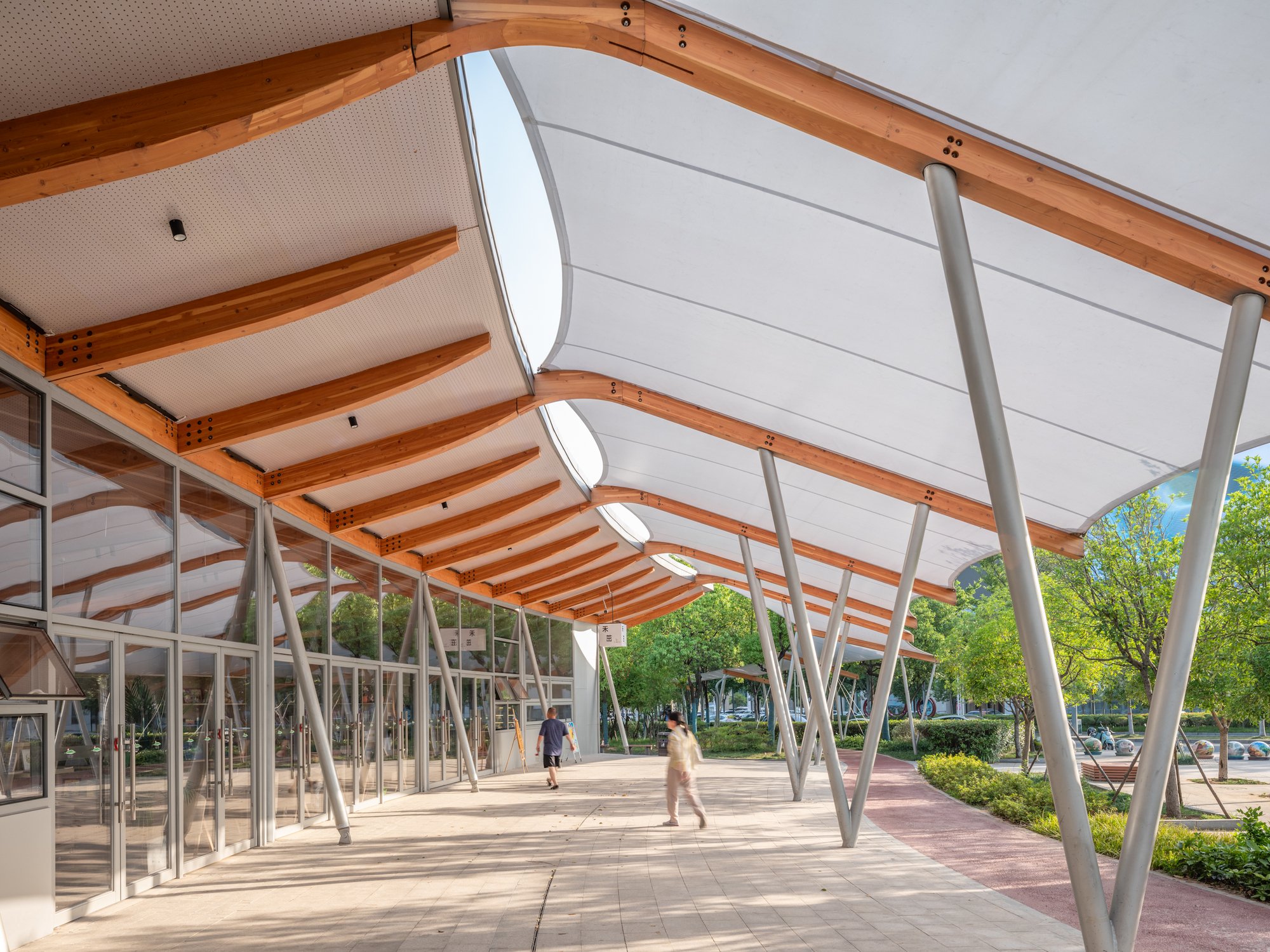 Walkway beneath the undulating canopy with timber ribs and tilted steel columns, two blurred pedestrians walking past a glazed frontage