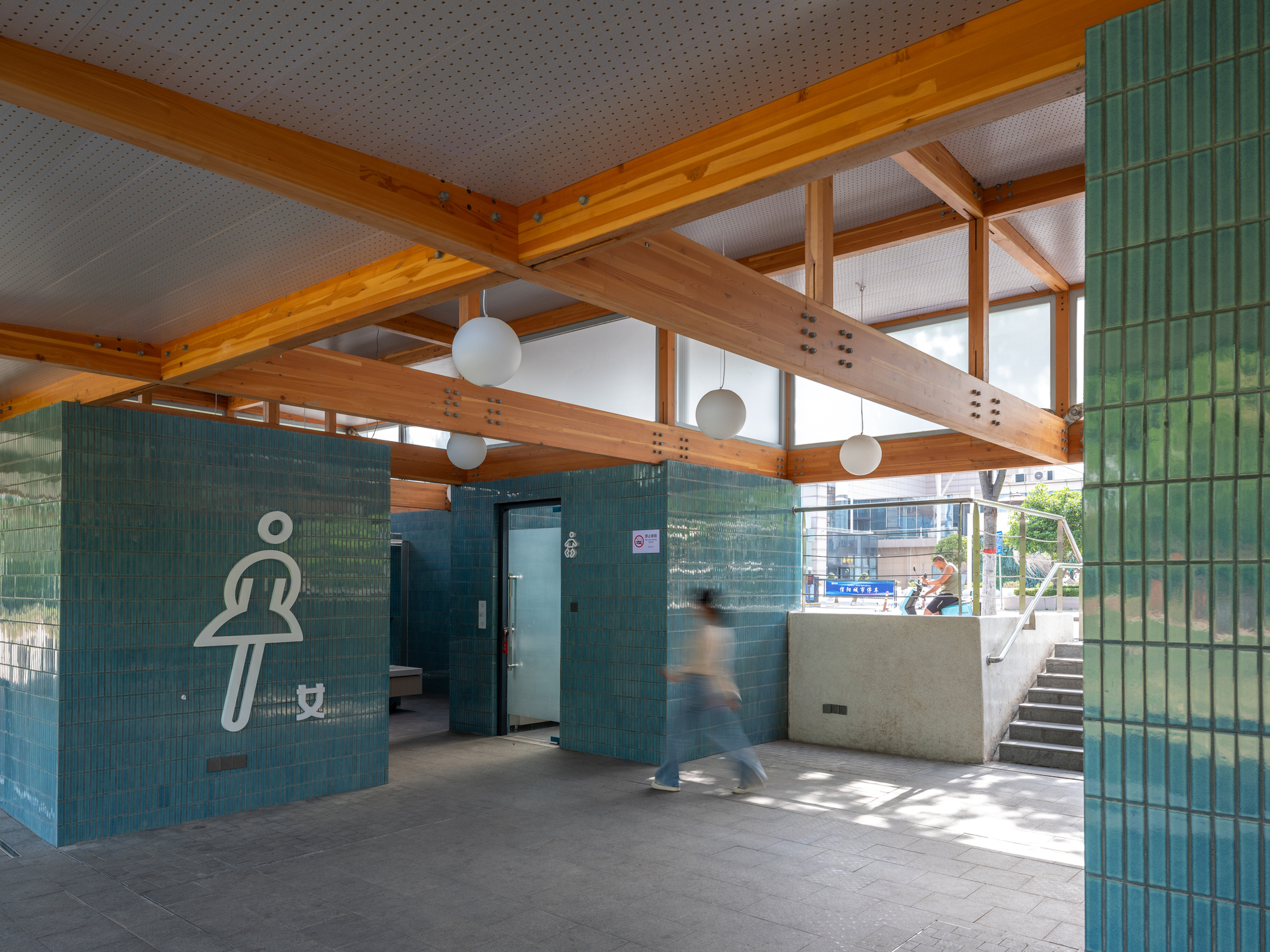 Teal tile restroom volumes under a glulam timber frame with globe lights, a blurred person crossing the paved floor