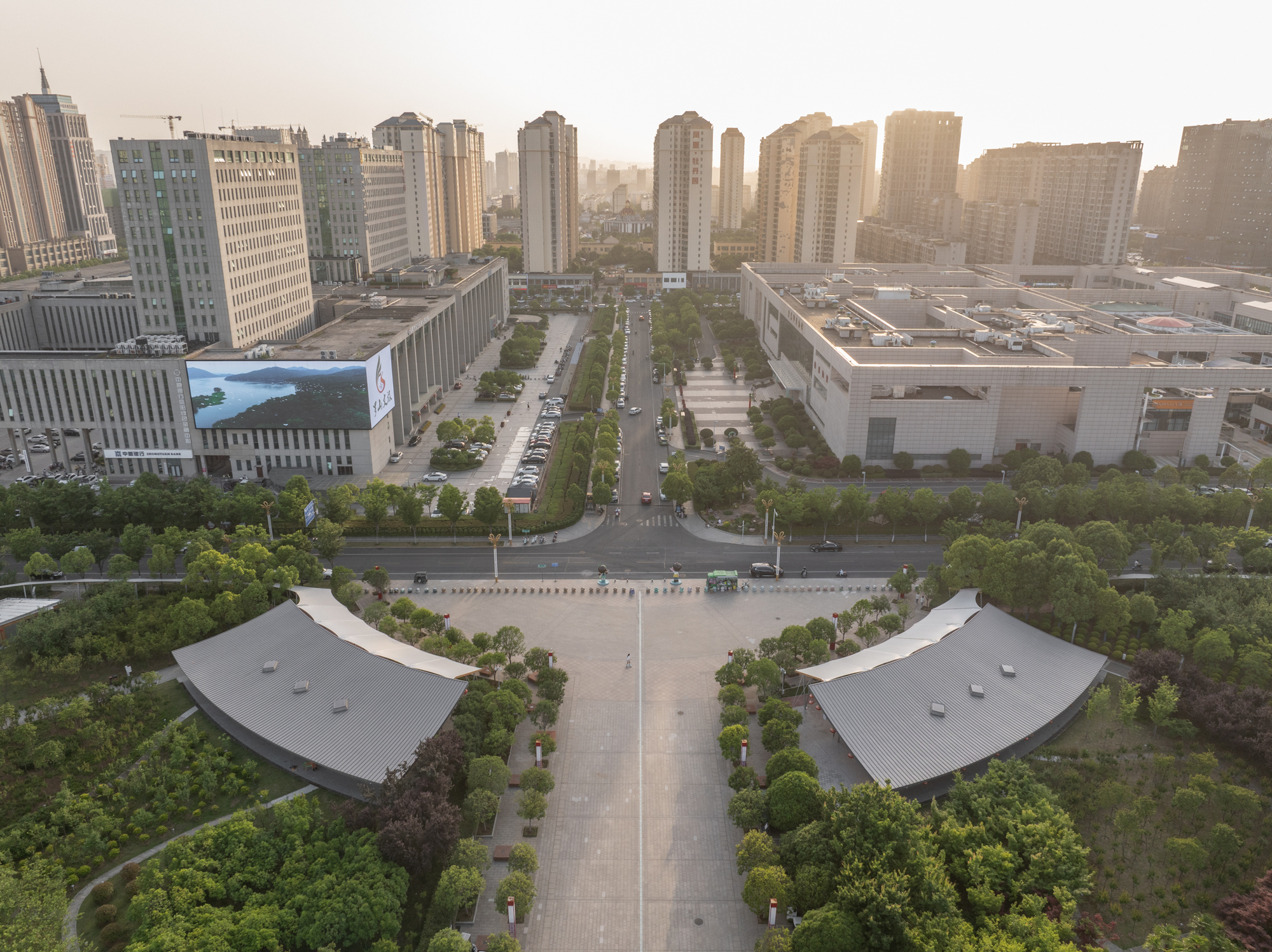 Aerial view of two curved metal-roofed pavilions flanking a paved axis toward high-rise towers in hazy evening light