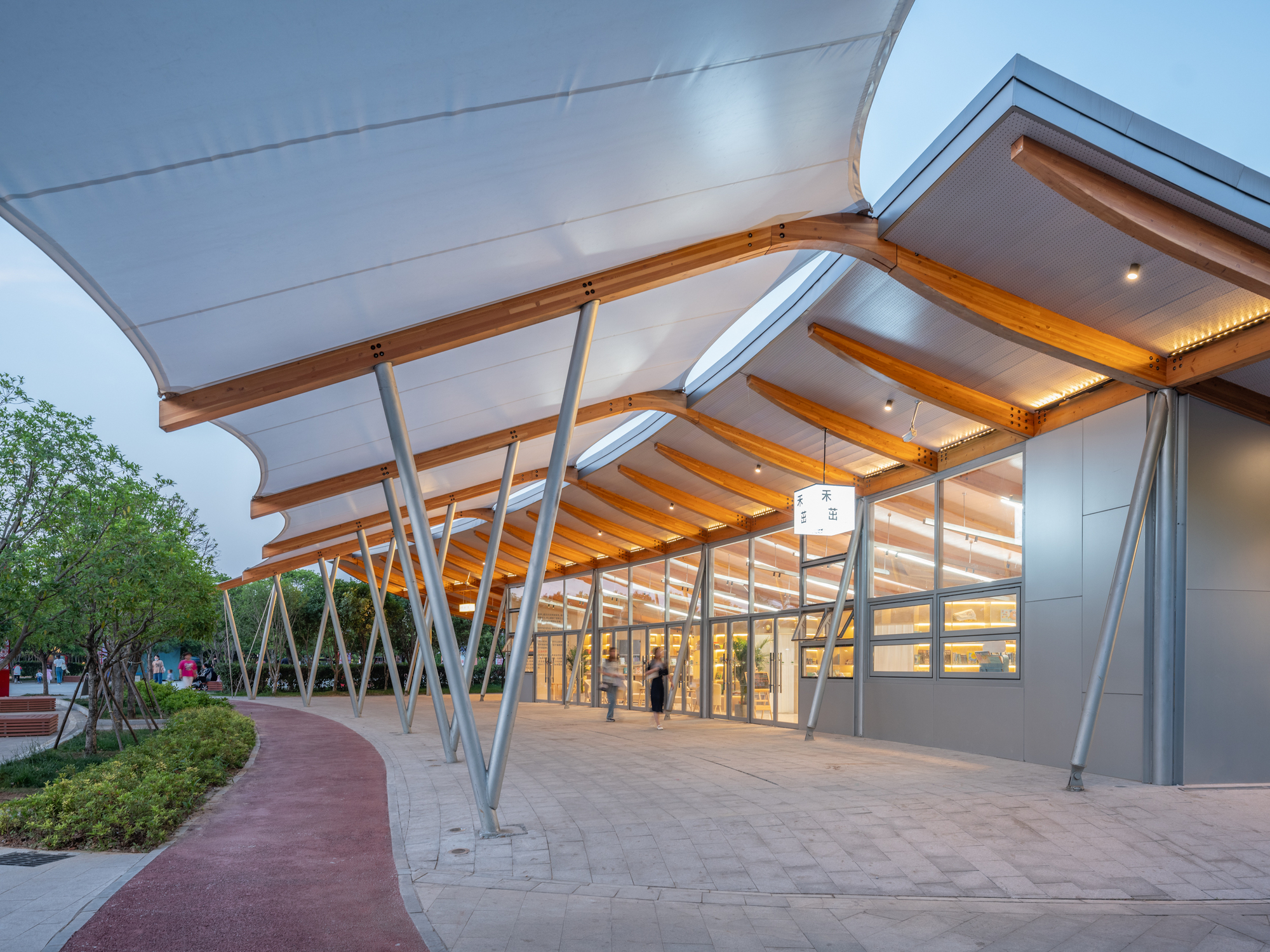 Tensile fabric canopy on slanted steel struts and timber ribs over a glazed pavilion, lit warmly at dusk