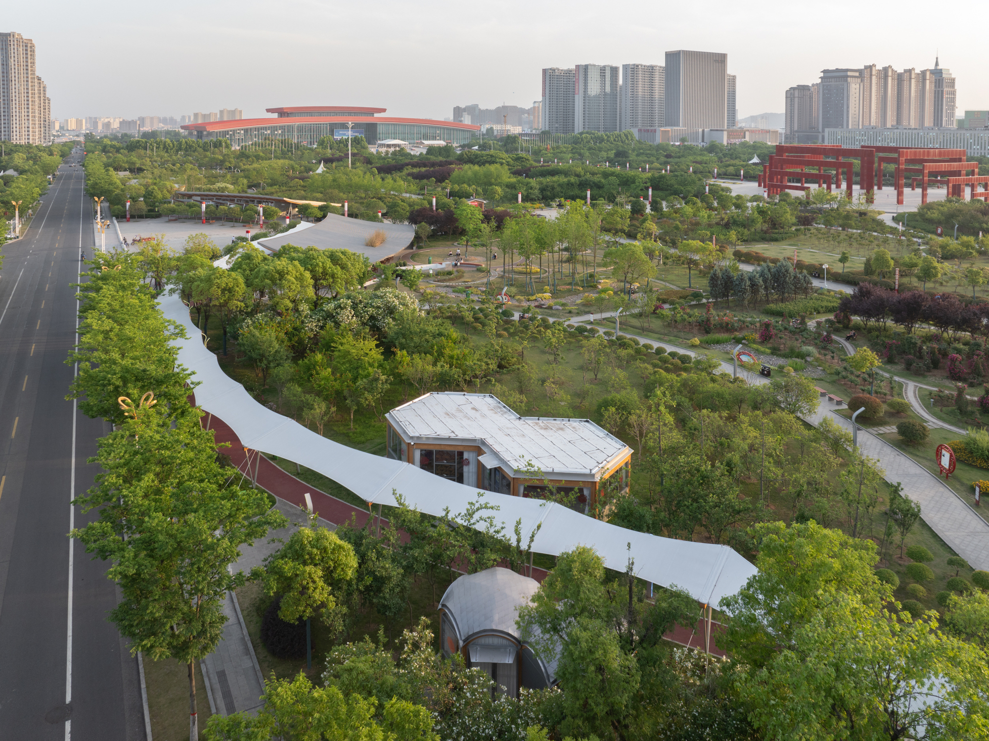 Aerial view of a long white fabric canopy winding through park trees past a small pavilion at sunset