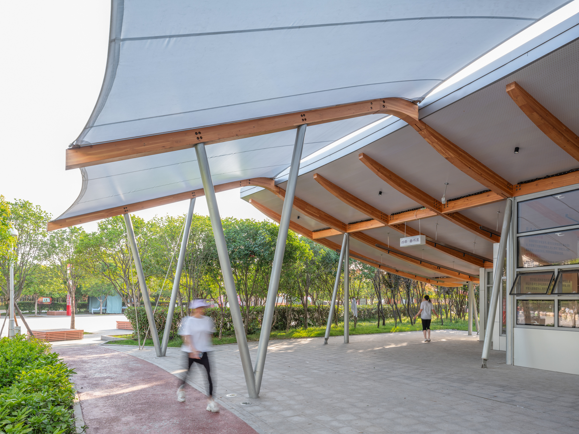 Fabric canopy edge and angled steel columns over a paved walkway with a blurred jogger passing in summer light