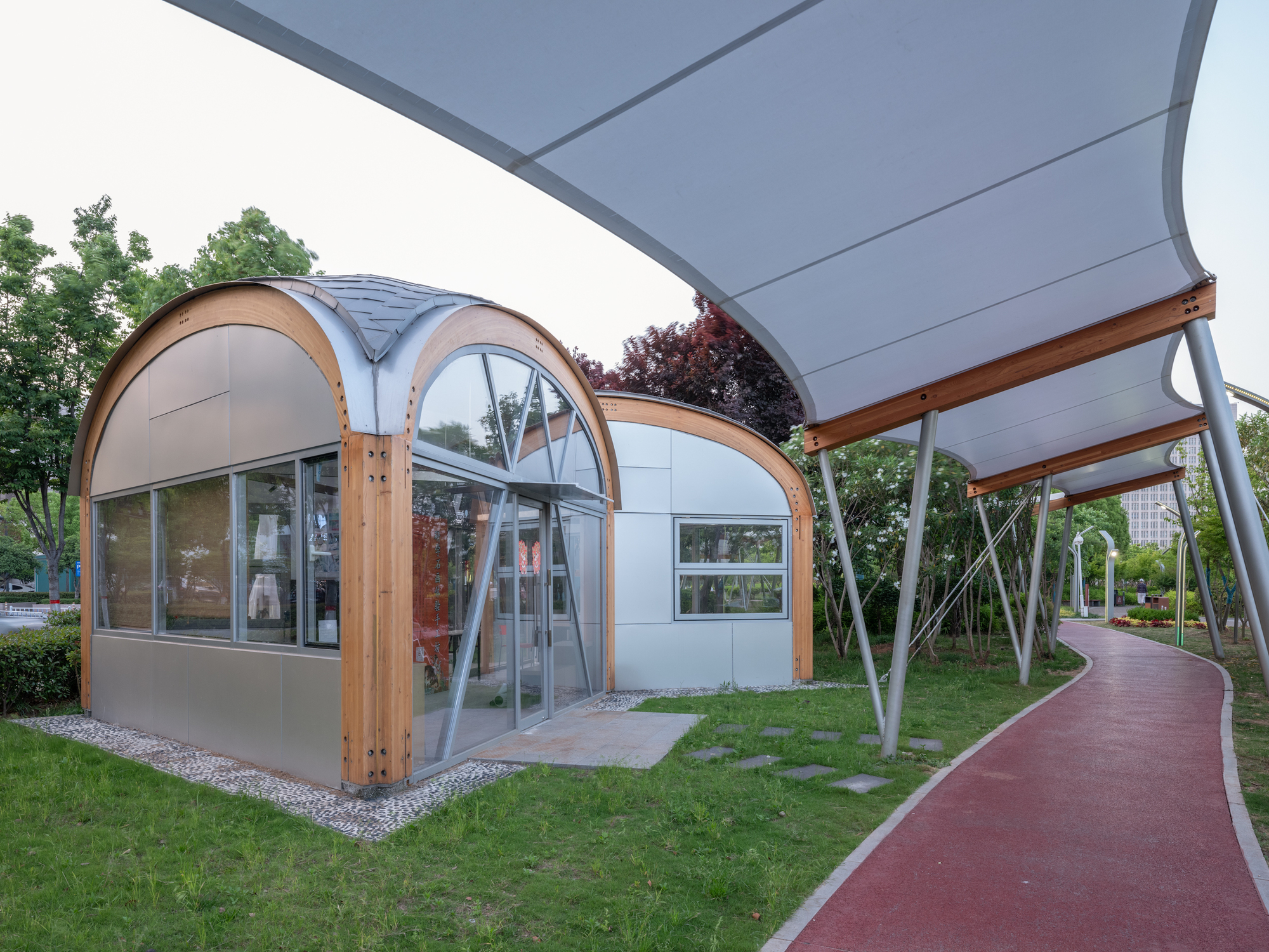 Barrel-vaulted timber-framed kiosk with glazed gable beside a red running path under the sweeping white canopy