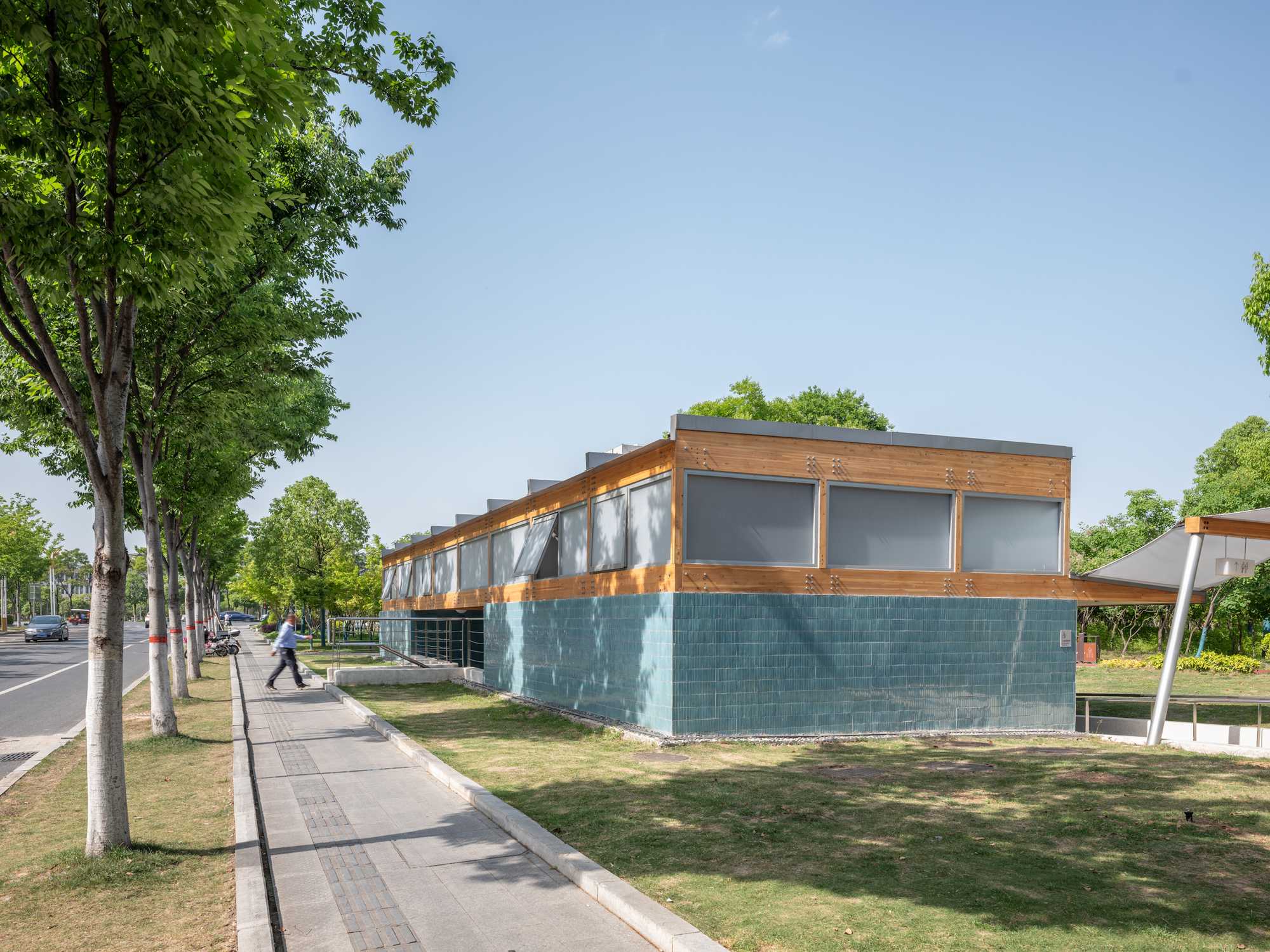 Low pavilion with teal glazed tile base and timber-framed upper band beside a sidewalk lined with young trees, under clear blue sky