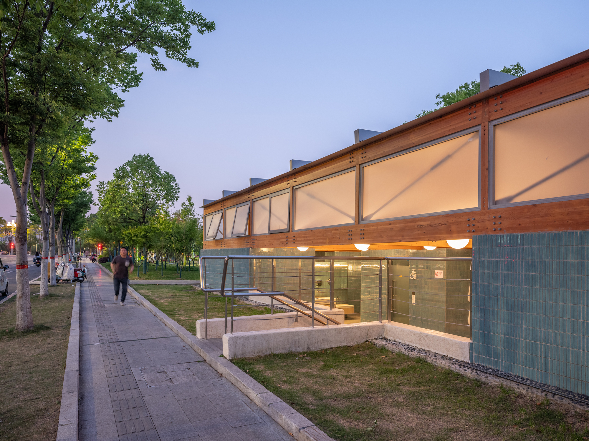 Dusk view of the pavilion's glowing translucent upper panels and sunken entry ramp with steel railings, a pedestrian walking past