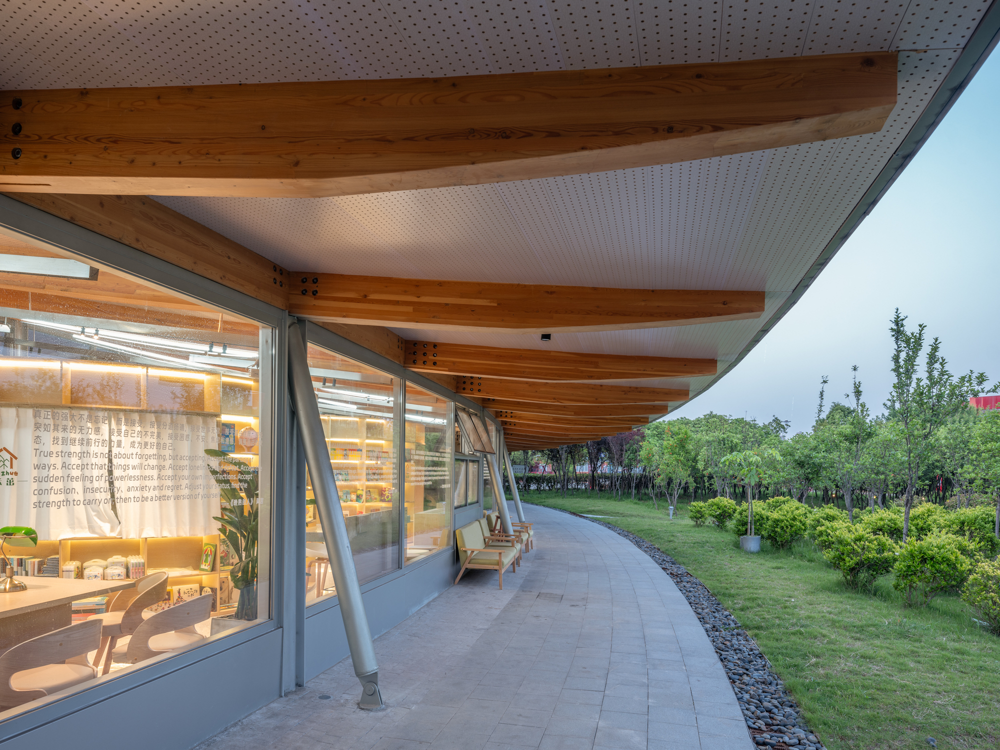 Curved timber-beamed overhang above a glazed reading room with slanted steel posts, a gravel edge and shrubs in evening light
