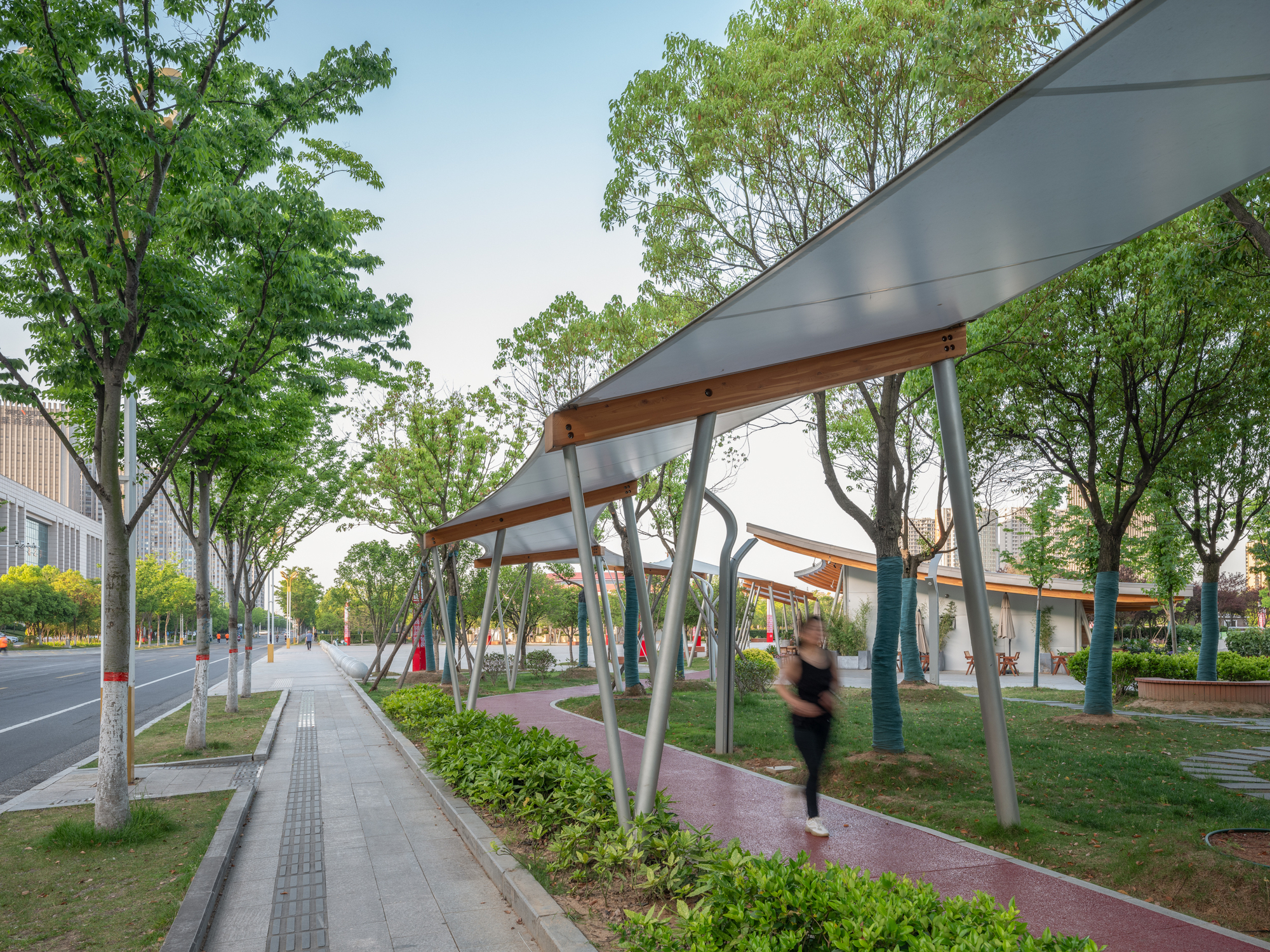 Row of tilted steel columns carrying translucent canopies with timber edges over a red path, a blurred walker passing on the pavement