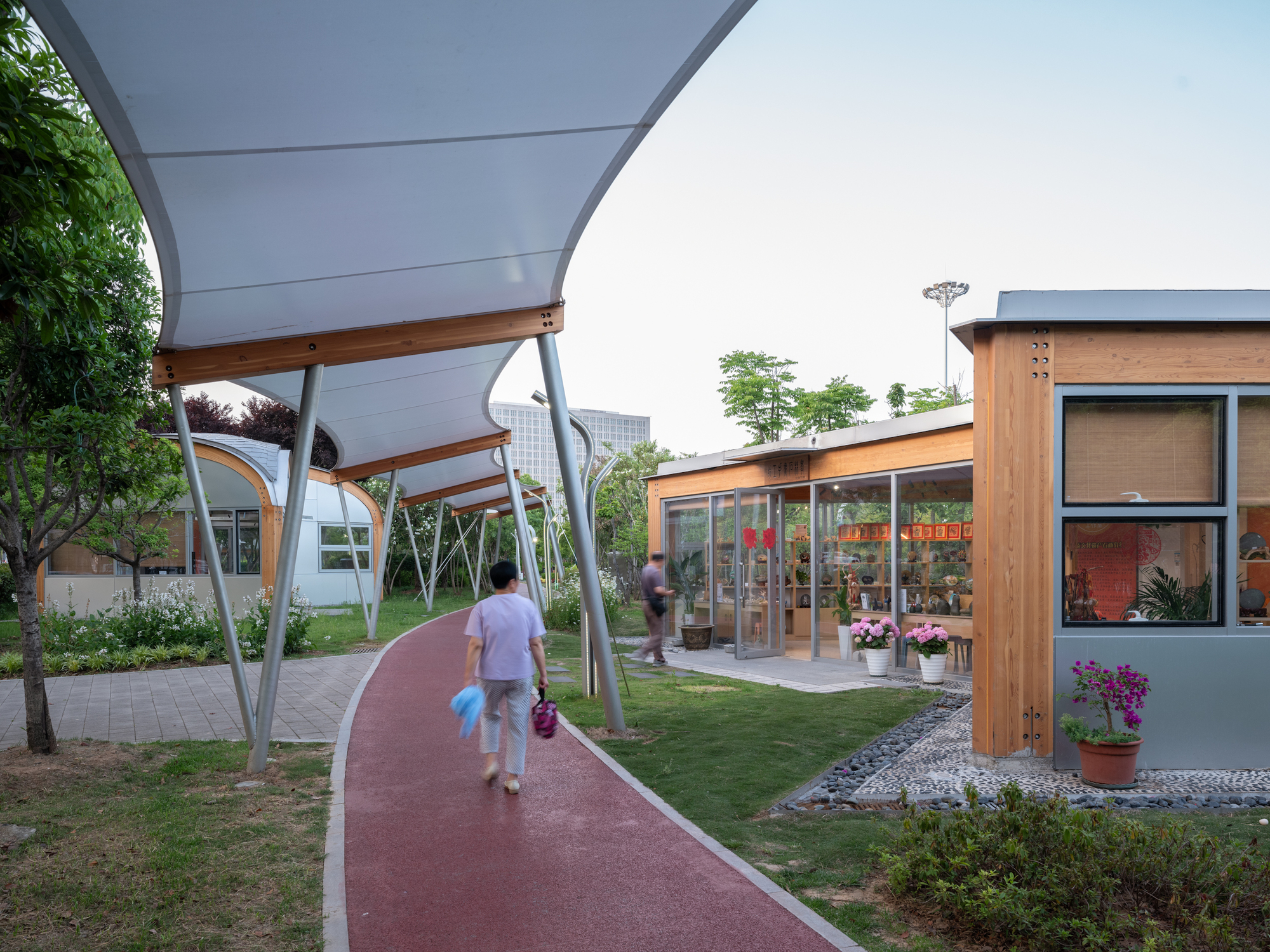Red walking path under a sweeping translucent canopy leading to timber-clad pavilions with glazed shopfronts and potted flowers, a woman strolling ahead