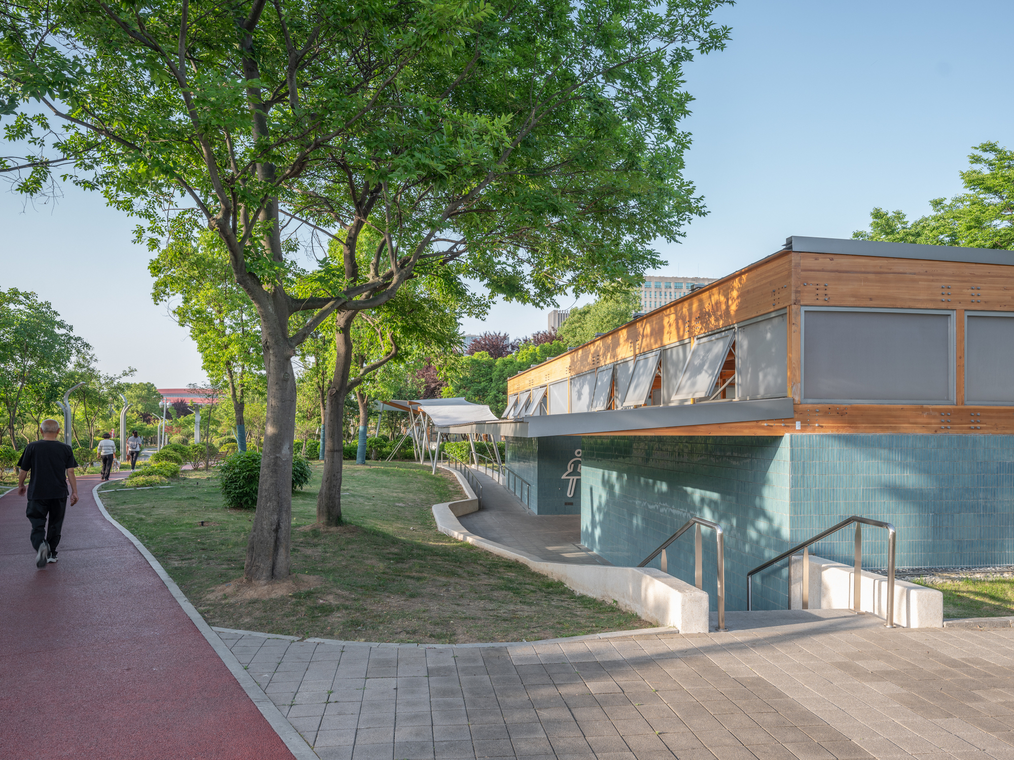 Timber-clad pavilion with teal tile base and shuttered windows beside a red running path, a pedestrian walking under park trees in afternoon sun