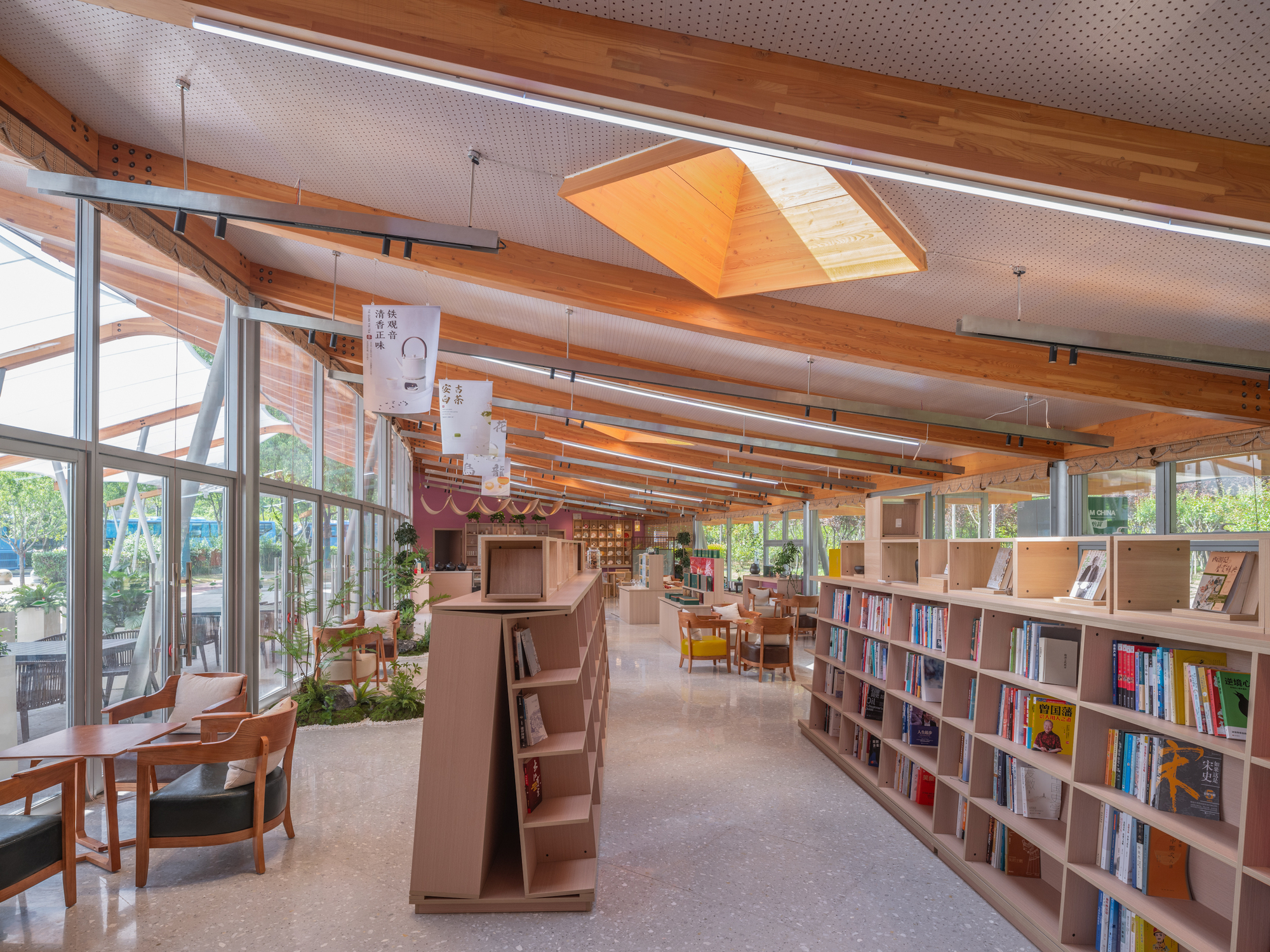Reading room with timber roof beams, a pyramidal skylight and wooden bookshelves running toward a glazed wall in diffuse daylight