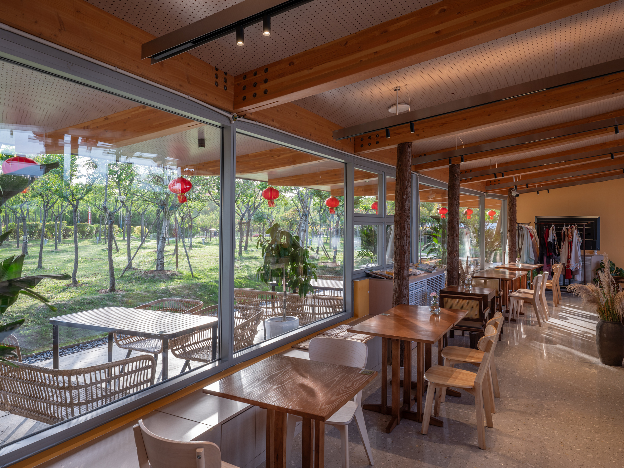 Cafe seating with wooden tables and a bark-clad column facing a glazed wall and red paper lanterns over a green lawn