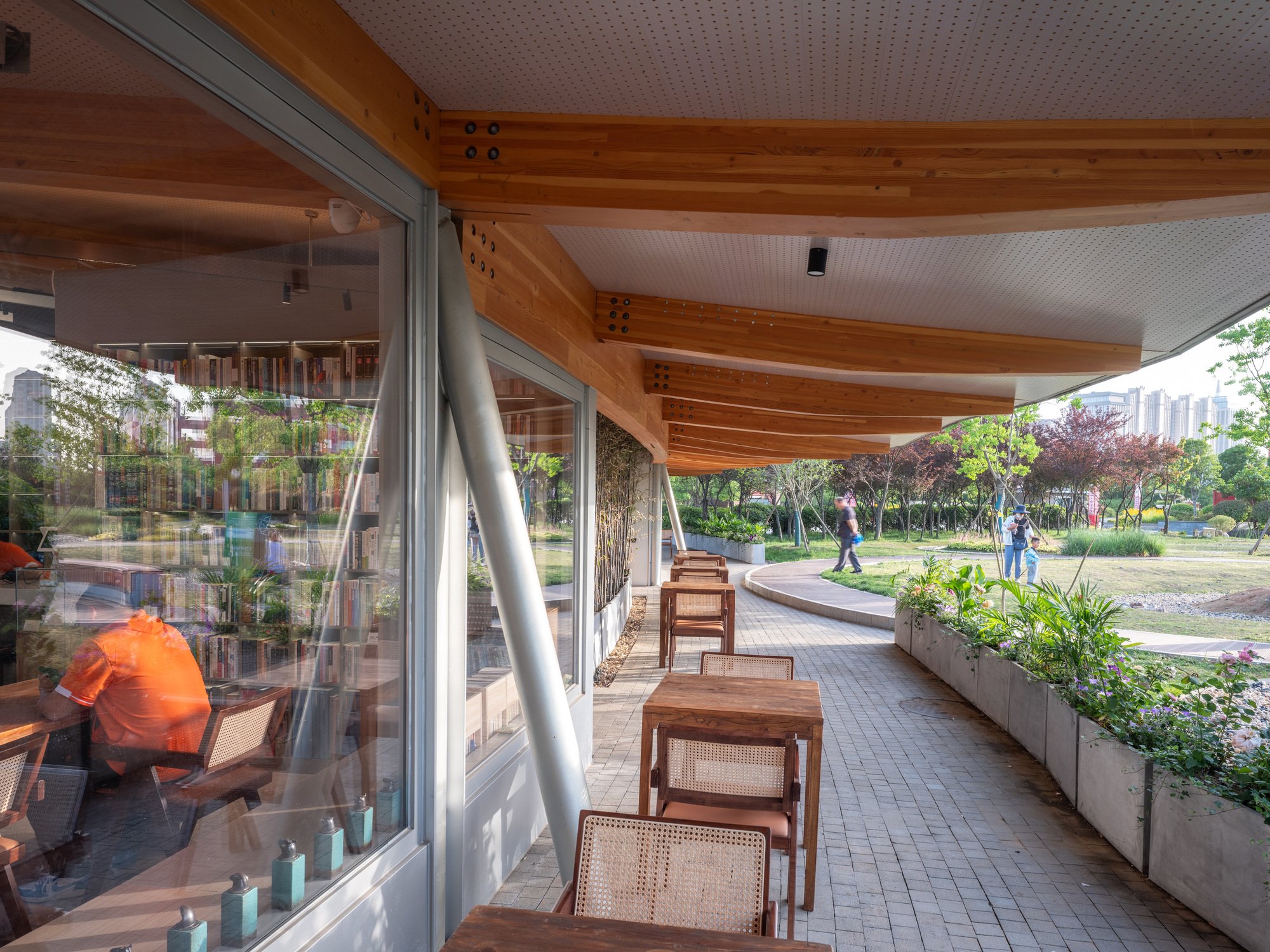 Covered walkway with timber beams, a slanted steel column and rattan chairs beside a planter bed, visitors strolling the park beyond