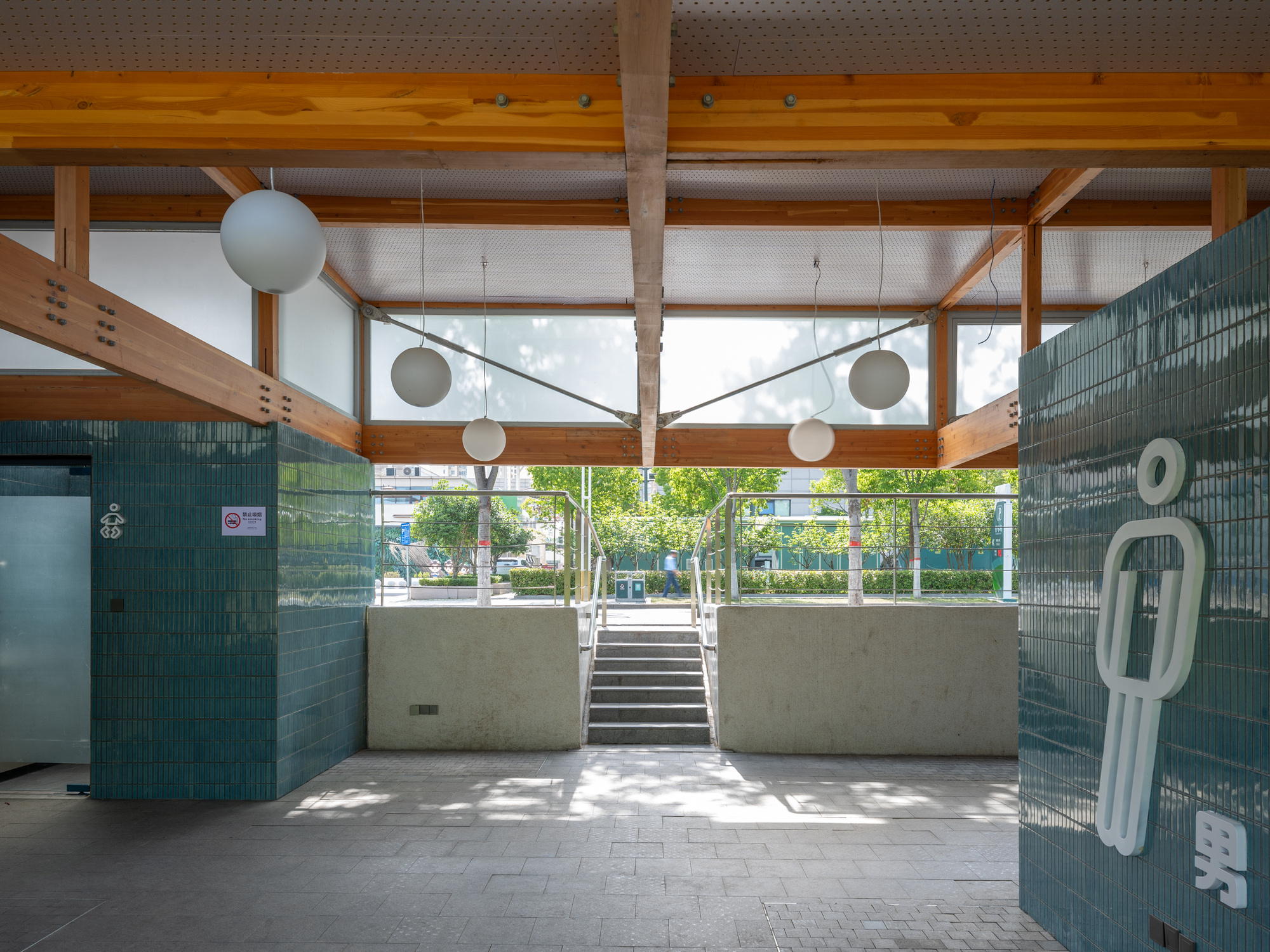 Restroom entrance hall with teal tile walls, a pictogram figure, spherical pendant lights and a stair rising to the park in daylight