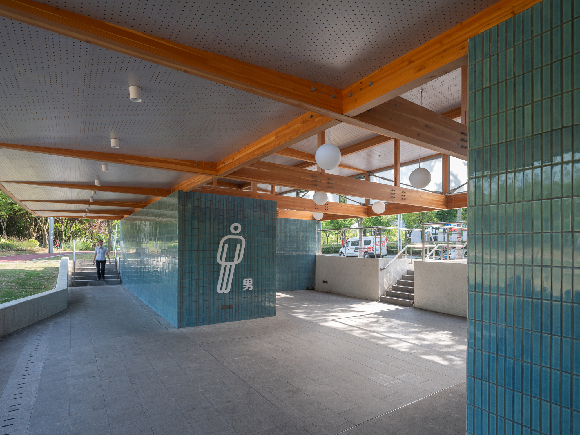 Open restroom entry with teal glazed tile walls, exposed timber beams and a pictogram sign, a visitor climbing steps in dappled daylight