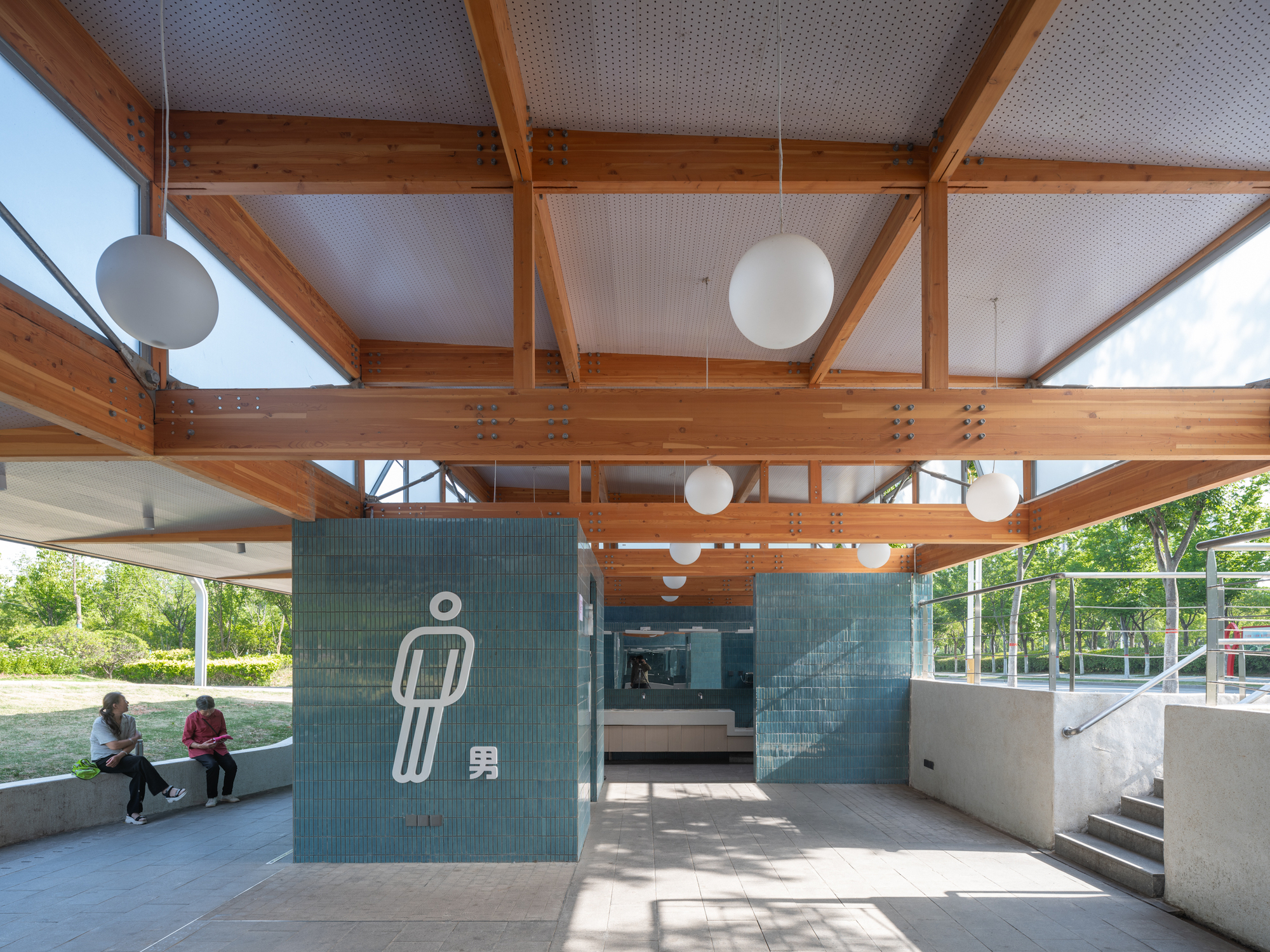 Timber truss ceiling with globe pendants above teal tile volumes, two people seated on a low concrete ledge in afternoon sun