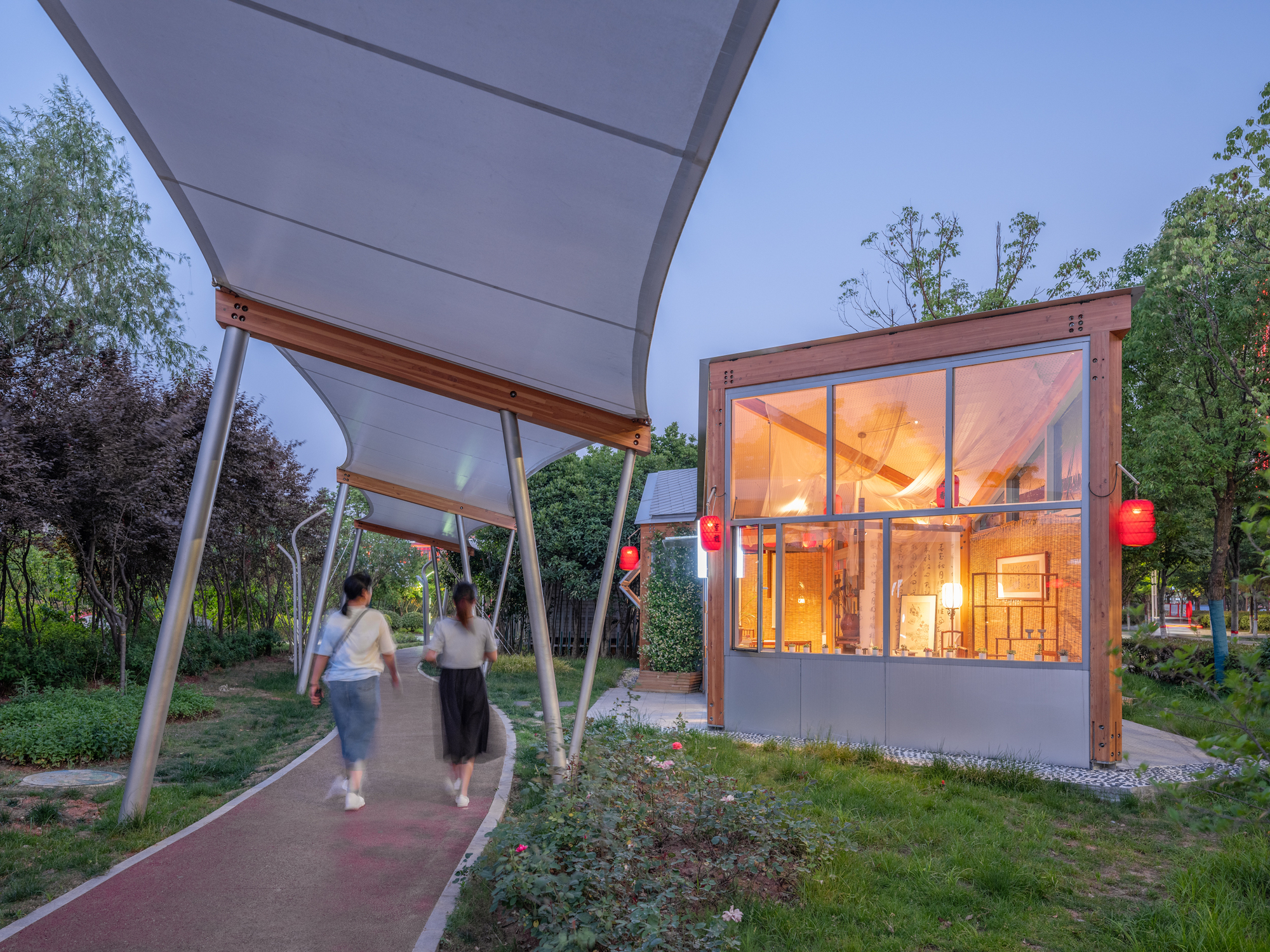 Two walkers pass under a curved fabric canopy toward a glowing timber-framed pavilion with red lanterns at dusk