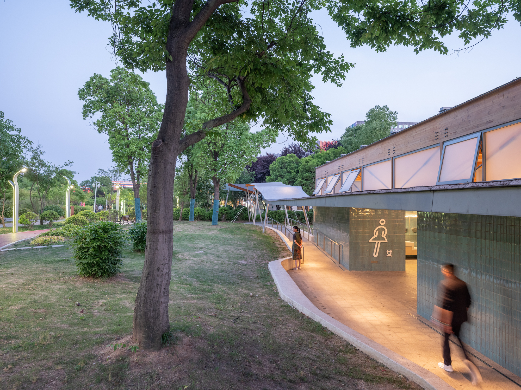 Teal tile restroom pavilion with a lit pictogram and clerestory windows, a blurred passerby on the lit path under dusk trees