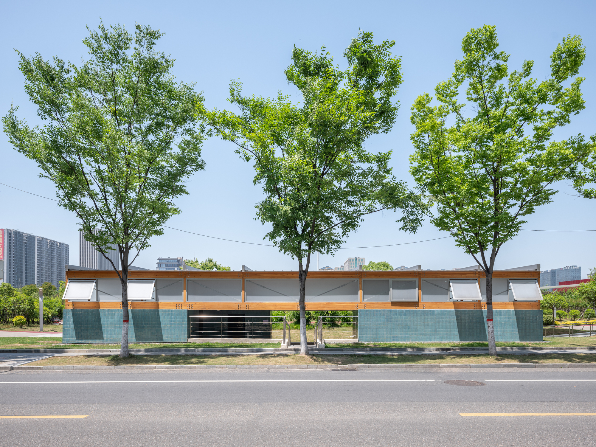 Street elevation of a low pavilion with teal tile base, timber band and awning windows behind three leafy trees in clear midday sun
