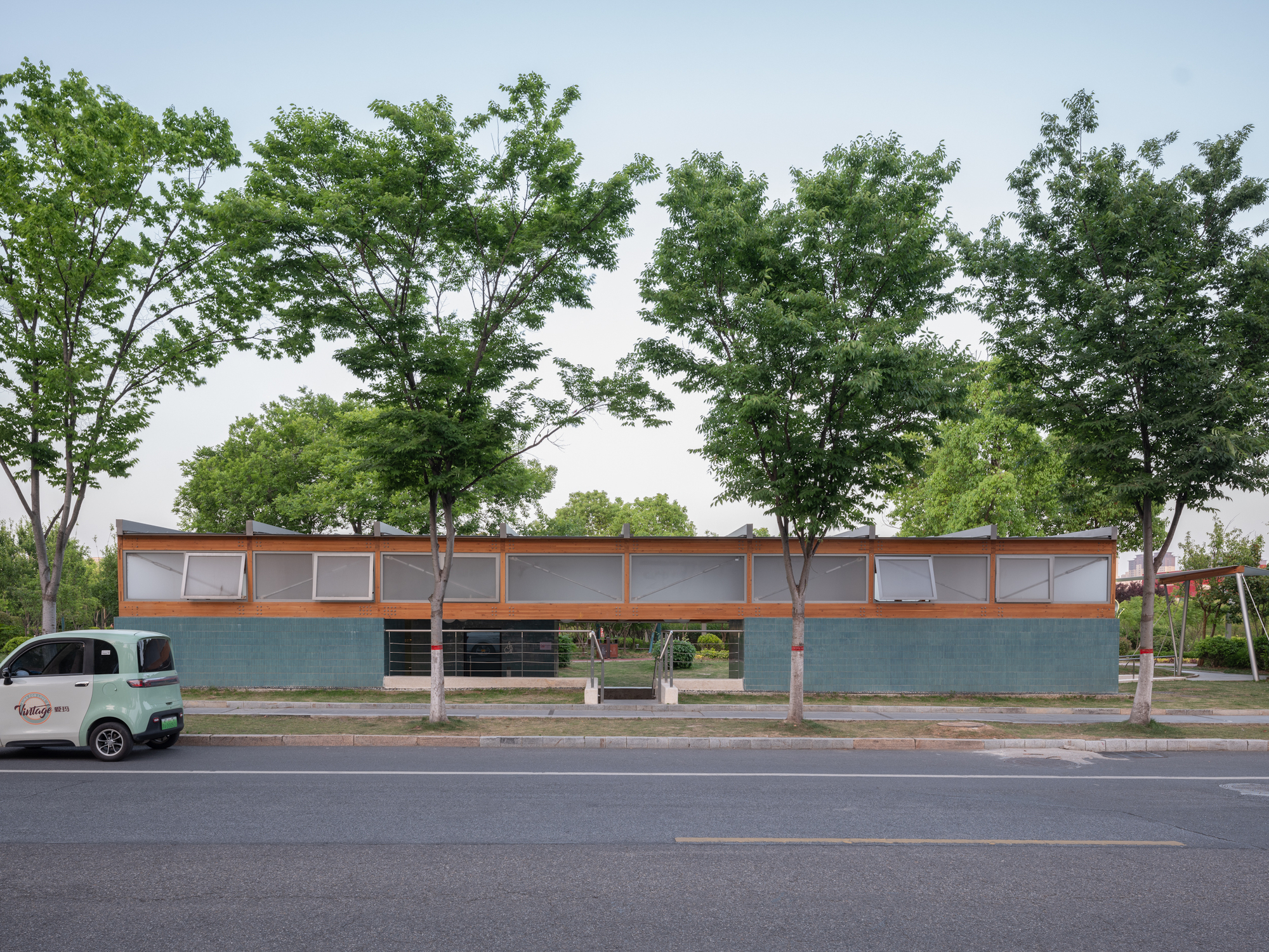 Long low pavilion with teal tiled walls and timber-framed clerestory windows, a small green car parked in front at dusk