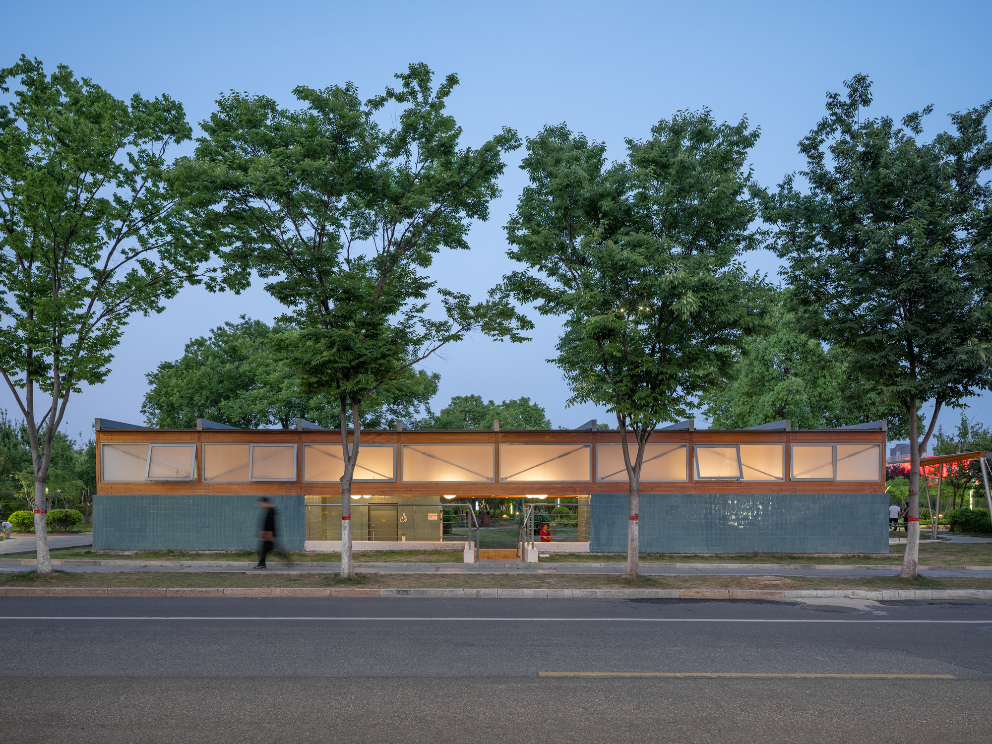 Pavilion glowing from within at twilight, its clerestory windows lit, with a blurred pedestrian walking past the street trees