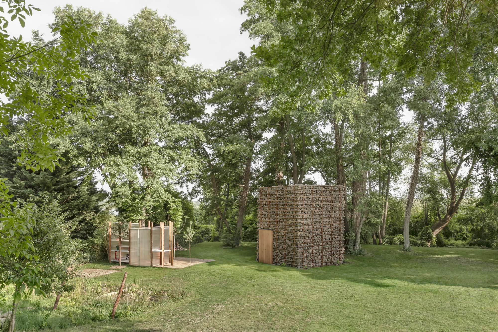 This photograph displays two small architectural pavilions nestled on an open grassy clearing surrounded by dense mature trees. One structure features a monolit