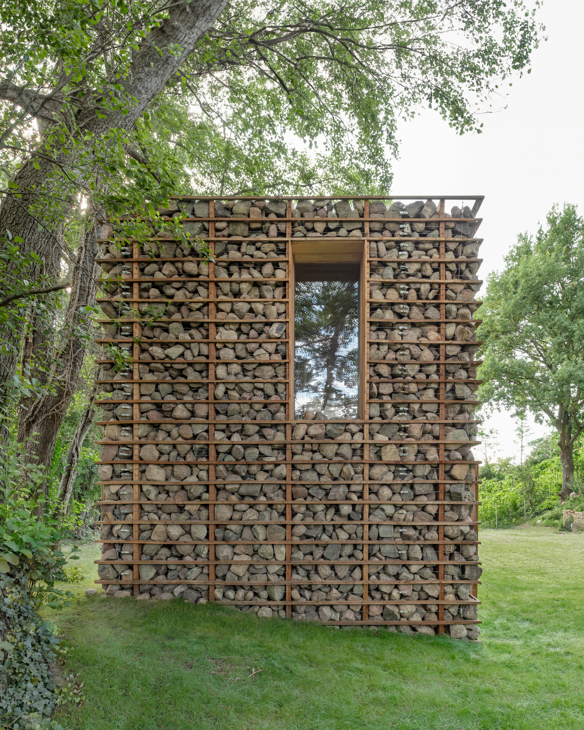 This photograph depicts a small, rustic modern building situated in a lush green landscape. The facade is uniquely constructed from dry-stacked river stones con