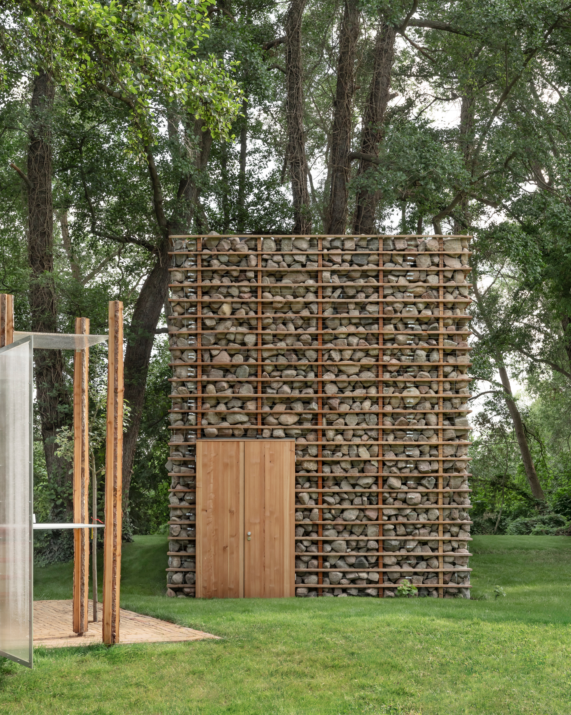 This outdoor photograph showcases a small architectural pavilion featuring heavy gabion walls filled with rounded natural fieldstones, retained by a grid of sle