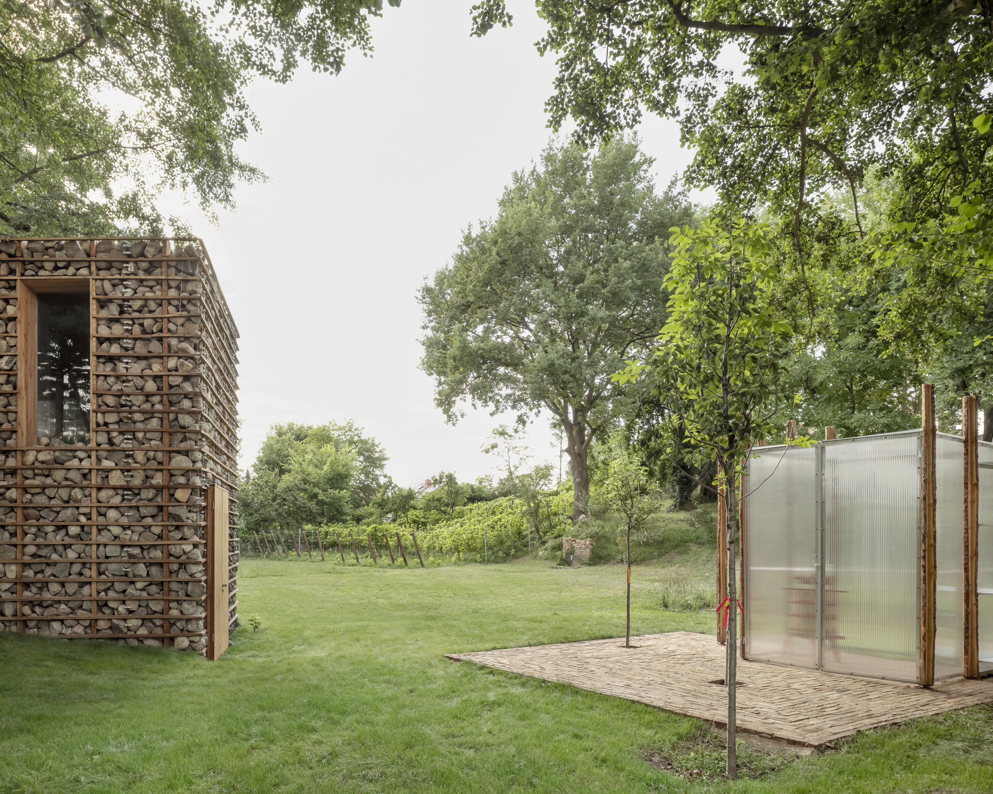 Stone-filled gabion tower beside a translucent polycarbonate pavilion on a brick paving pad, with vineyard rows and oak trees behind