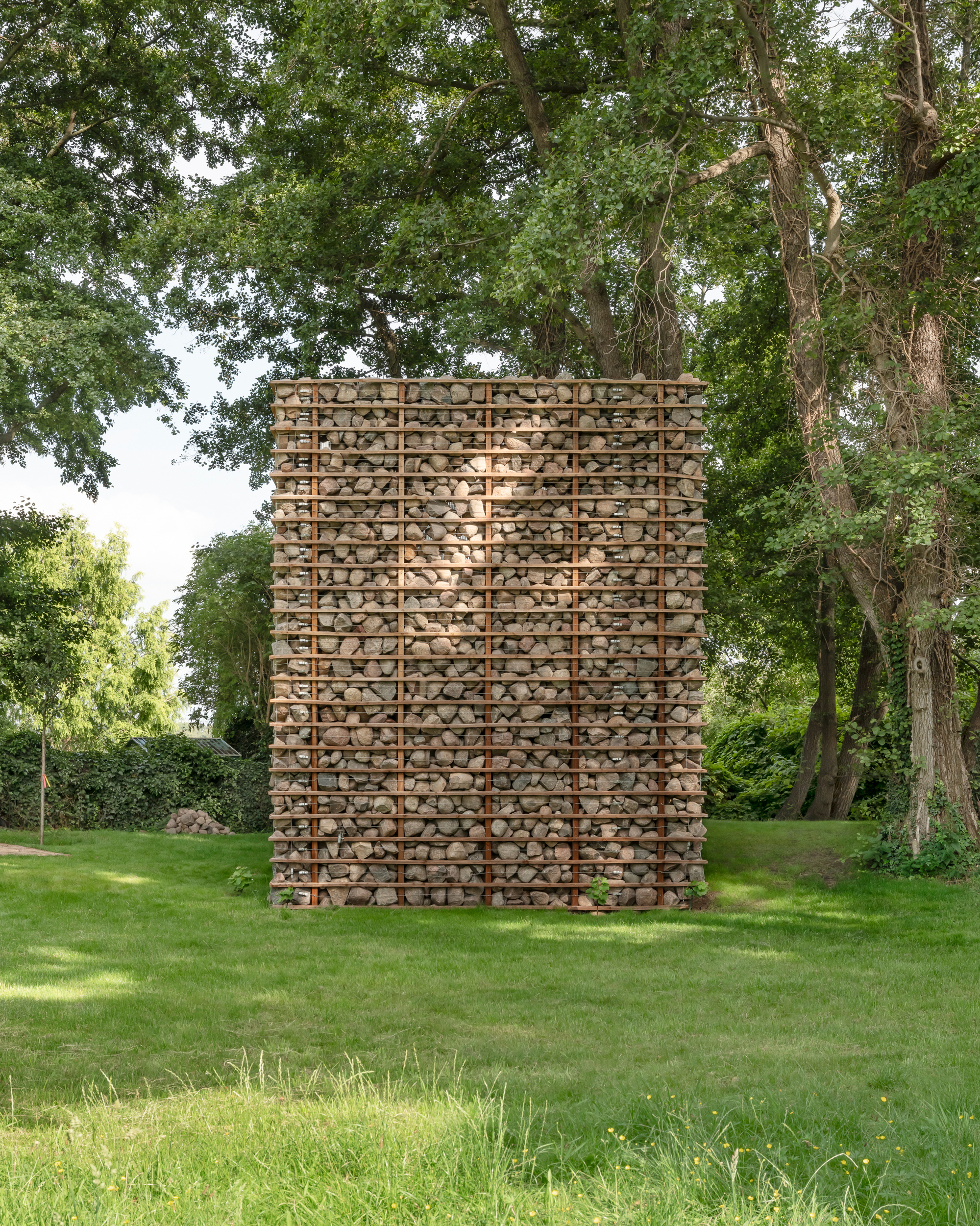 Freestanding stone-filled tower framed by horizontal rusted steel bars, standing on a lawn beneath tall trees in summer light