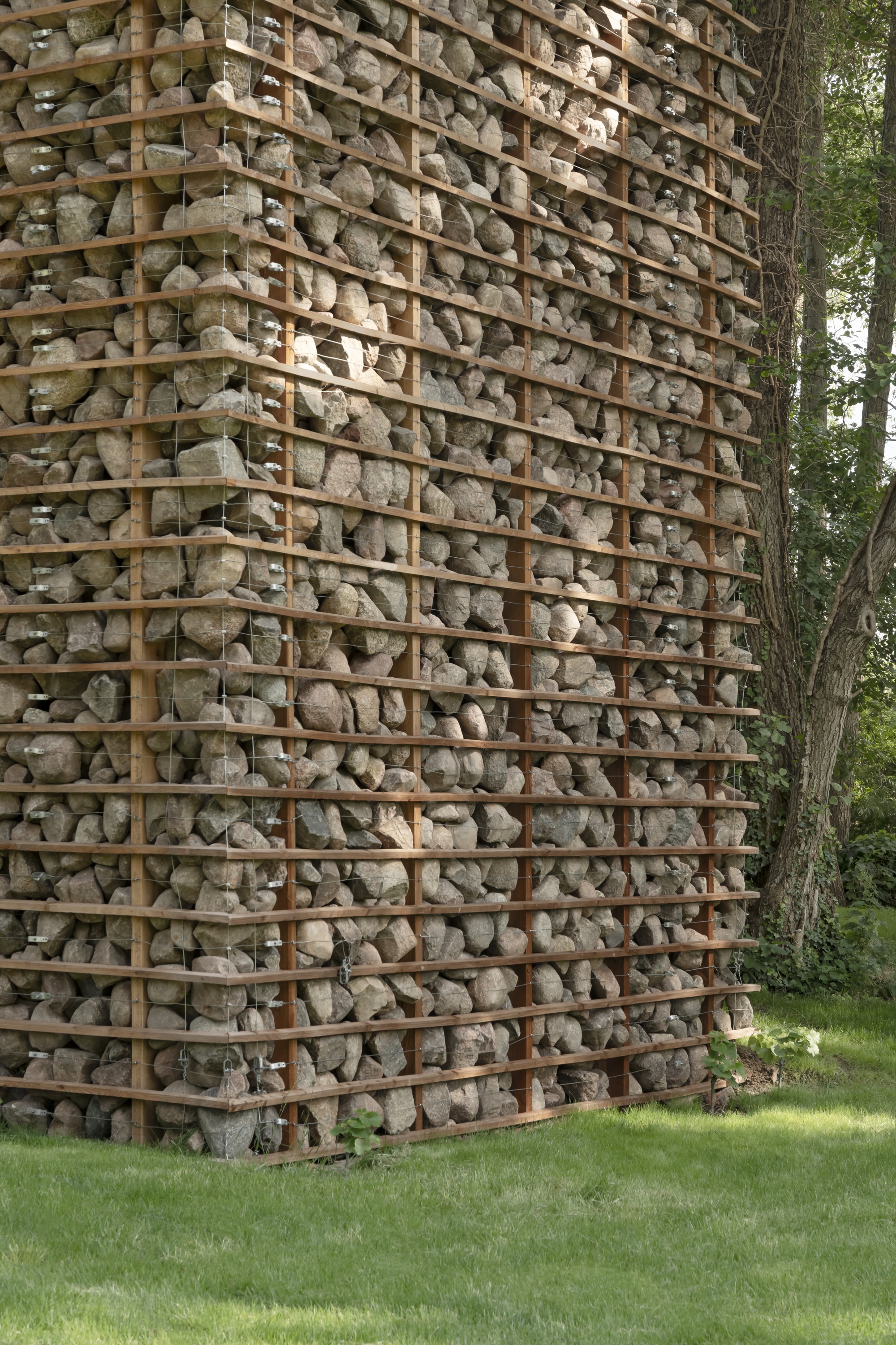Close-up of the tower corner showing fieldstones held between steel flats and wire mesh, with a tree trunk beside it