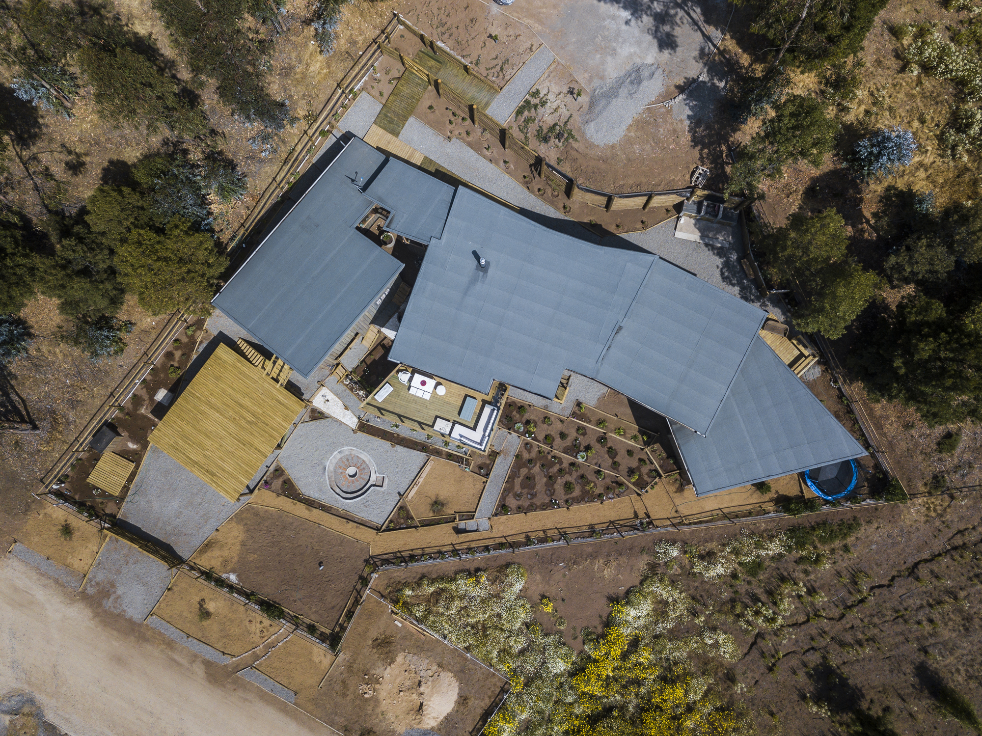 Drone view of the angled grey metal roofs, timber decks and terraced garden beds on a dry scrubby hillside