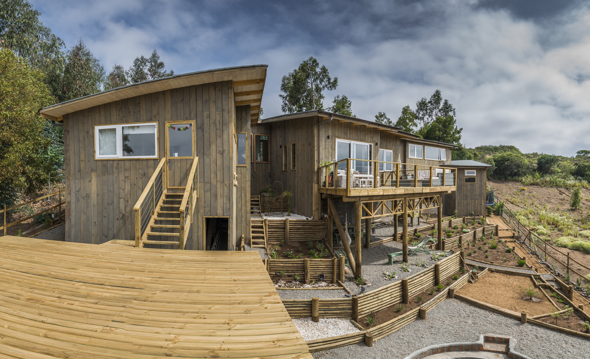 Timber-clad volumes with a raised deck and stepped retaining beds under a cloudy sky with eucalyptus behind