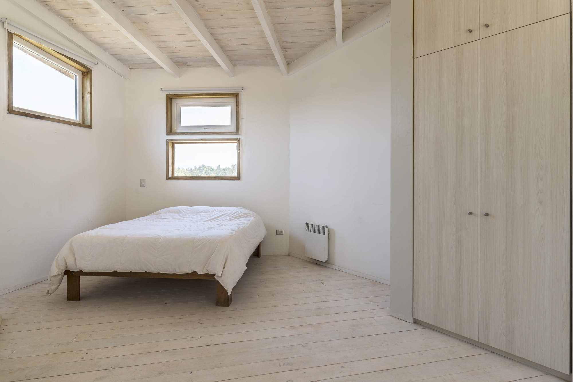 Bedroom with a low wooden bed, whitewashed plank floor and pale wardrobe doors beside small timber-framed windows