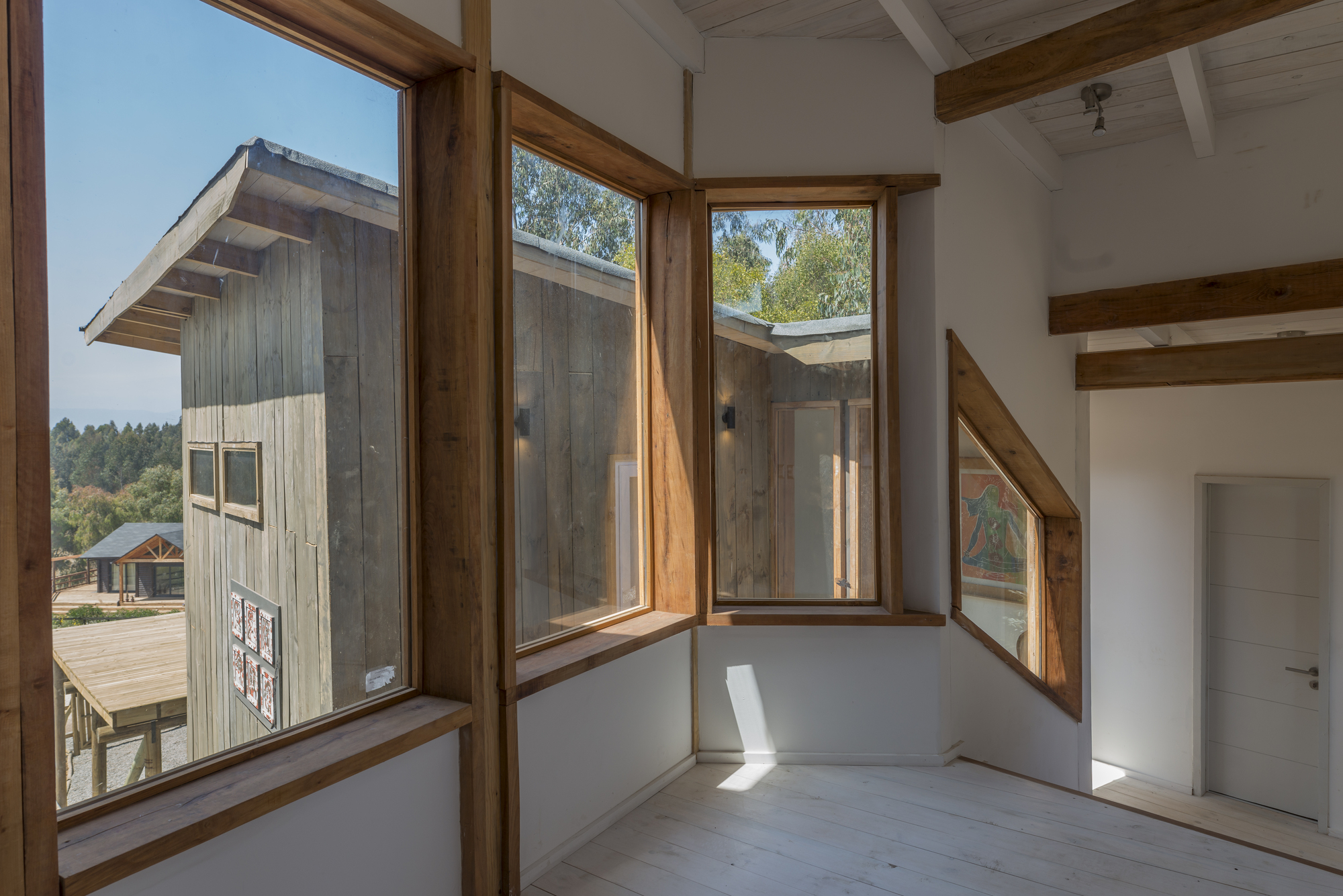 Corner of an upper room with timber-framed windows, one diagonal, looking onto a plank-clad wing in bright sun