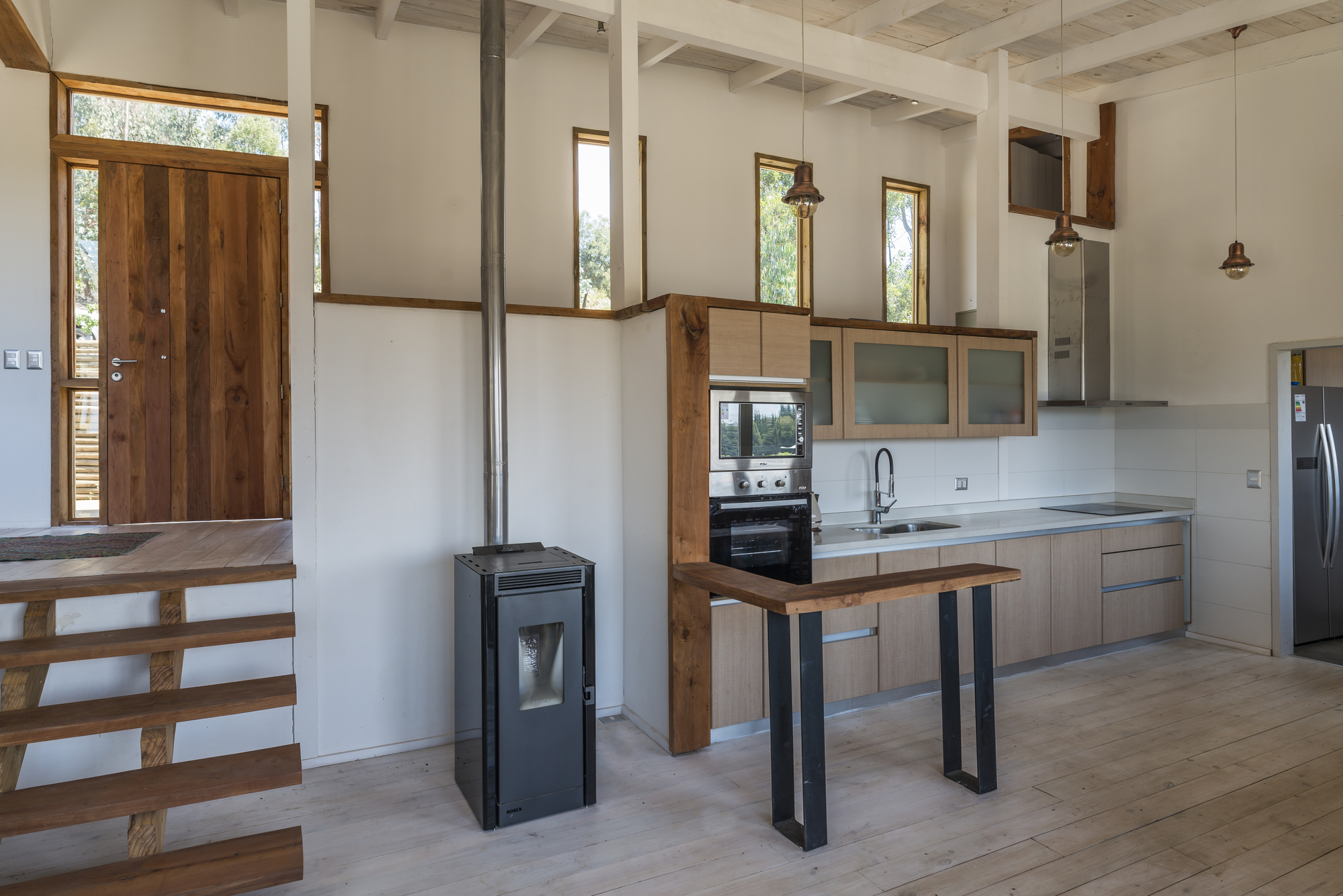 Open kitchen with a pellet stove, steel-legged timber counter and tall narrow windows above a wooden stair, lit by afternoon sun