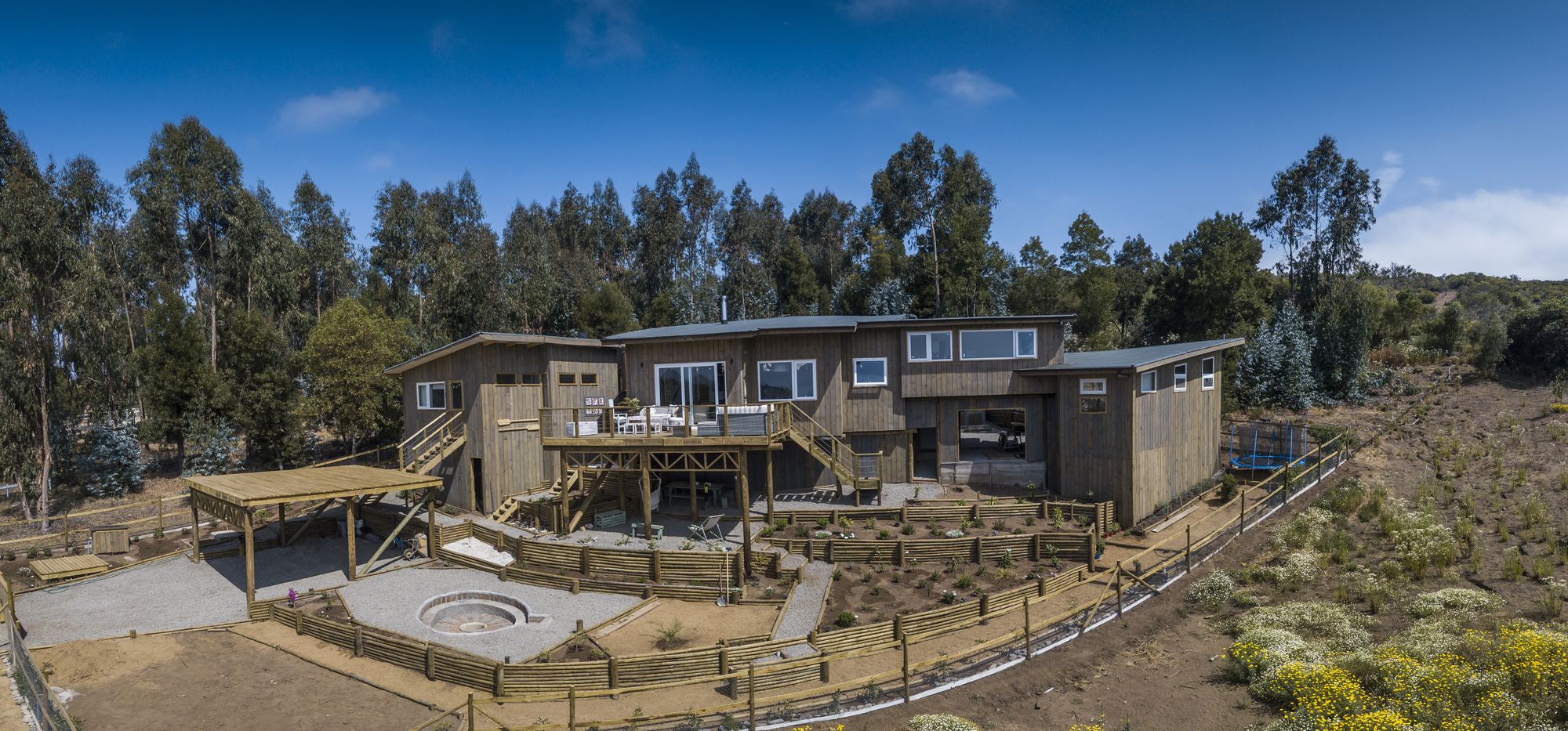 Wide view of the timber-clad house with raised deck, stepped garden beds and fire pit below a blue sky with eucalyptus behind