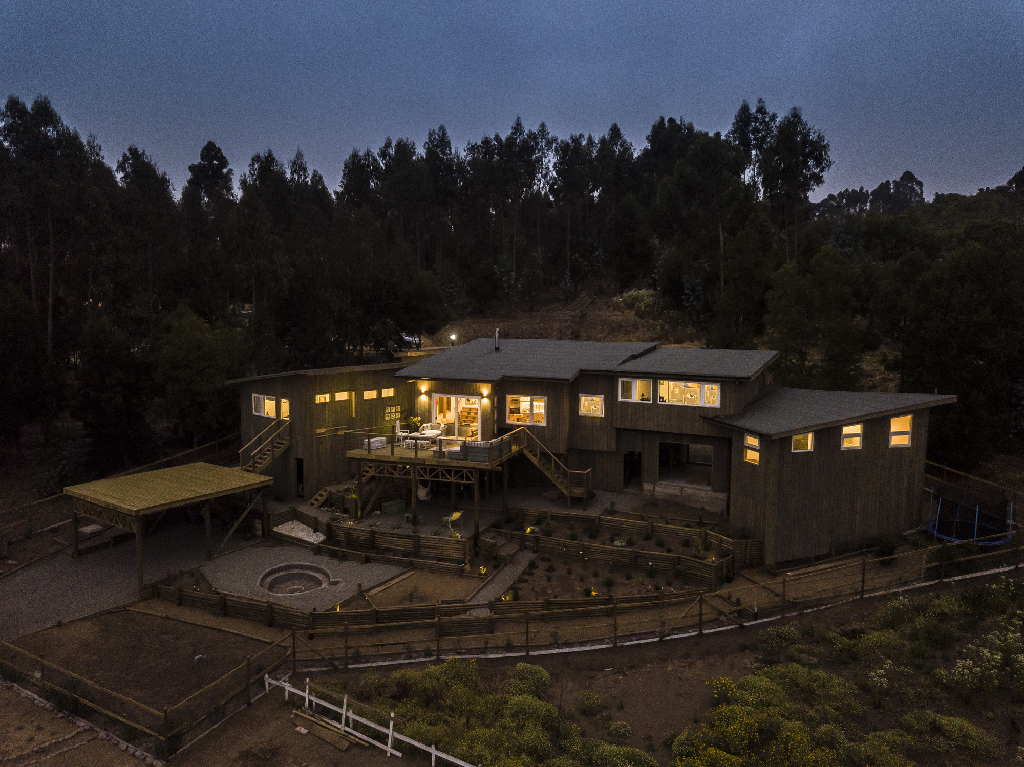 Aerial view of the lit timber house at dusk, with warm windows, deck stairs and a fenced fire pit terrace