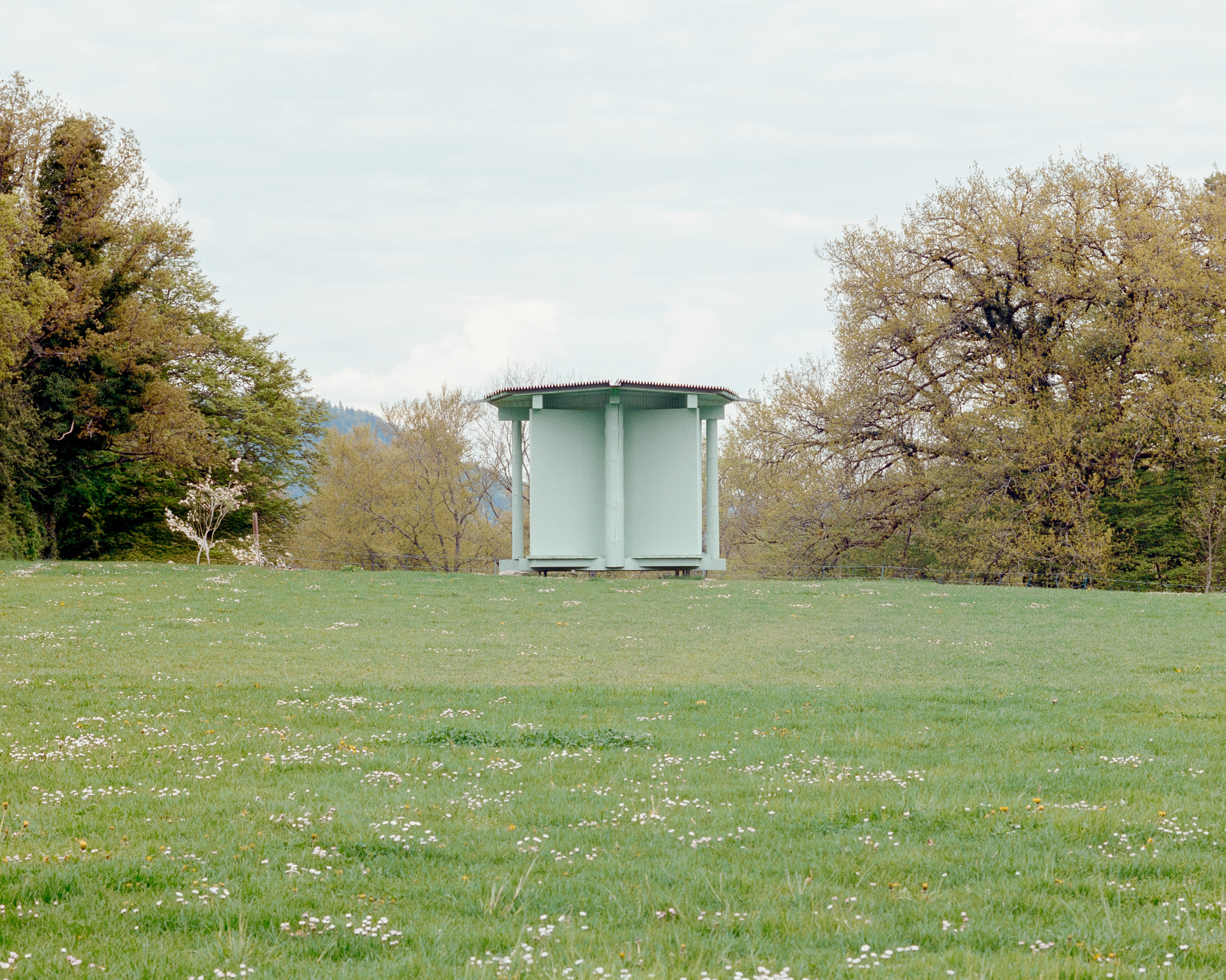 Small pale green pavilion with closed solid panels on a raised platform, set in a daisy-dotted meadow under overcast sky