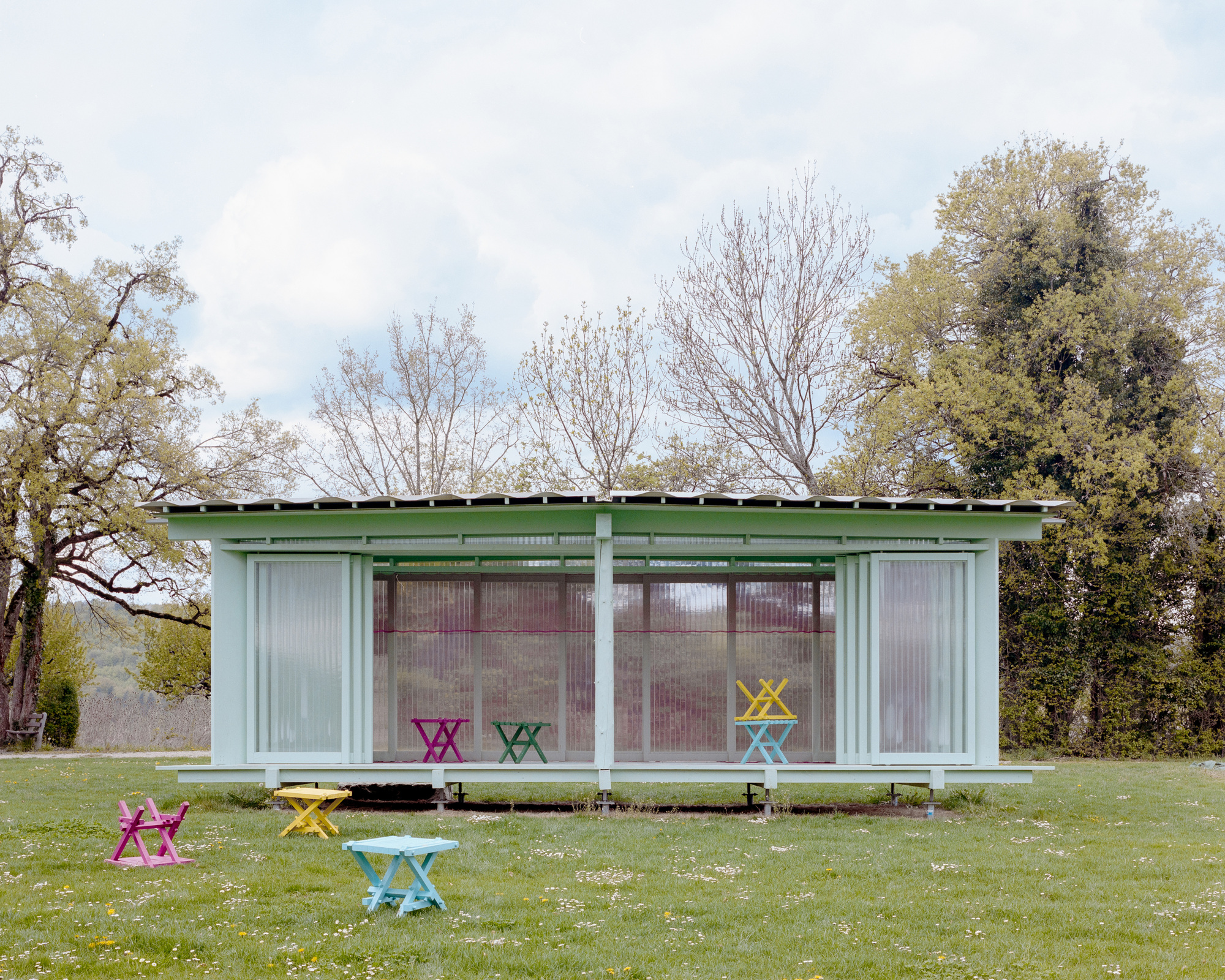 Open-sided pale green pavilion with translucent sliding panels and brightly colored folding stools scattered on the lawn in spring