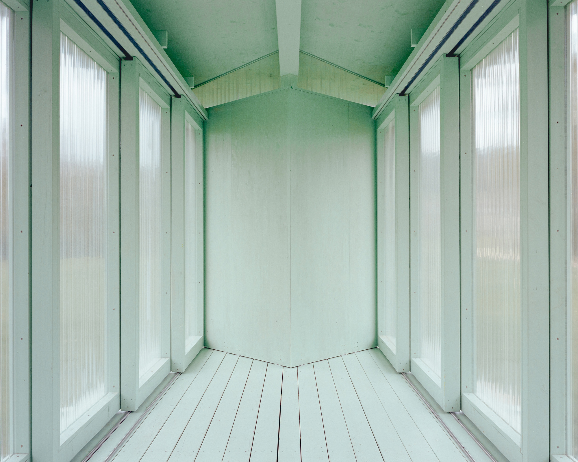 Interior corridor of the pavilion with fluted translucent panels, painted floorboards and an angled end wall in soft green
