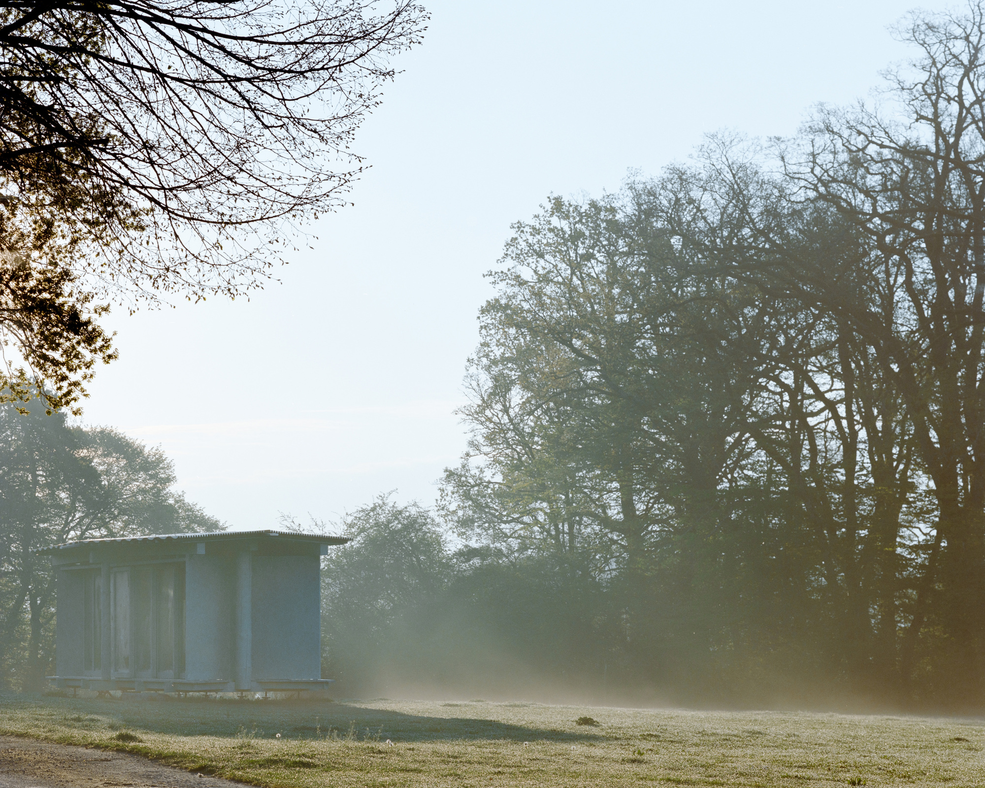 Pavilion with corrugated roof emerging from morning mist and backlit oak trees at the edge of a frosted field