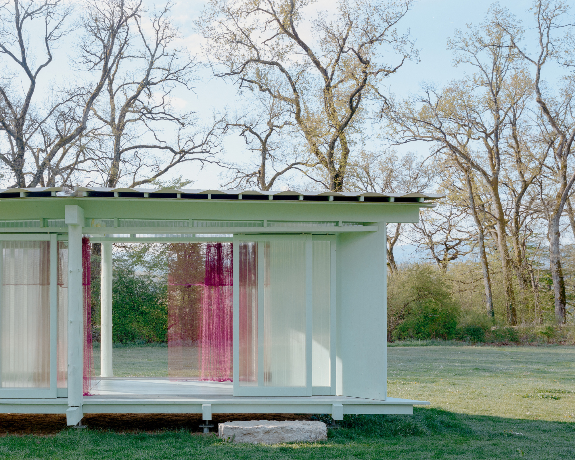 Corner of a pale green pavilion with sliding translucent panels and magenta curtains, framed by bare oaks in spring light