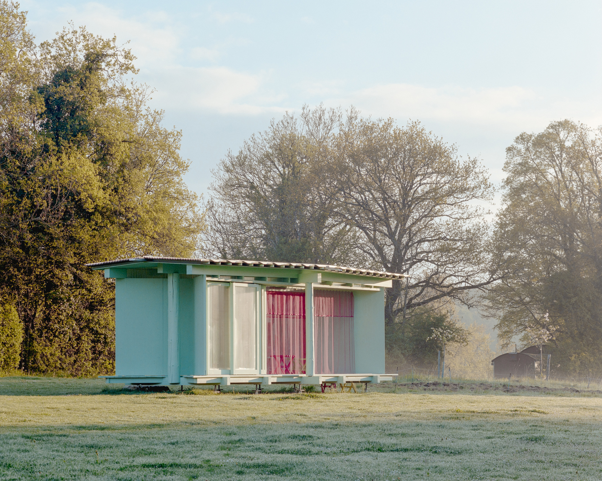 Small raised pavilion with a corrugated roof and pink curtains standing on frosted grass in early morning haze