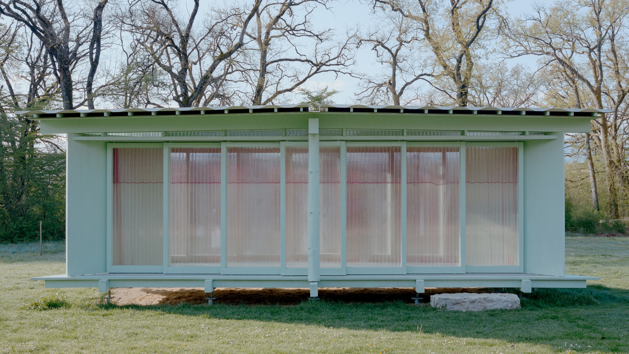 Straight-on view of the glazed pavilion front with a central post and a limestone block resting on the lawn