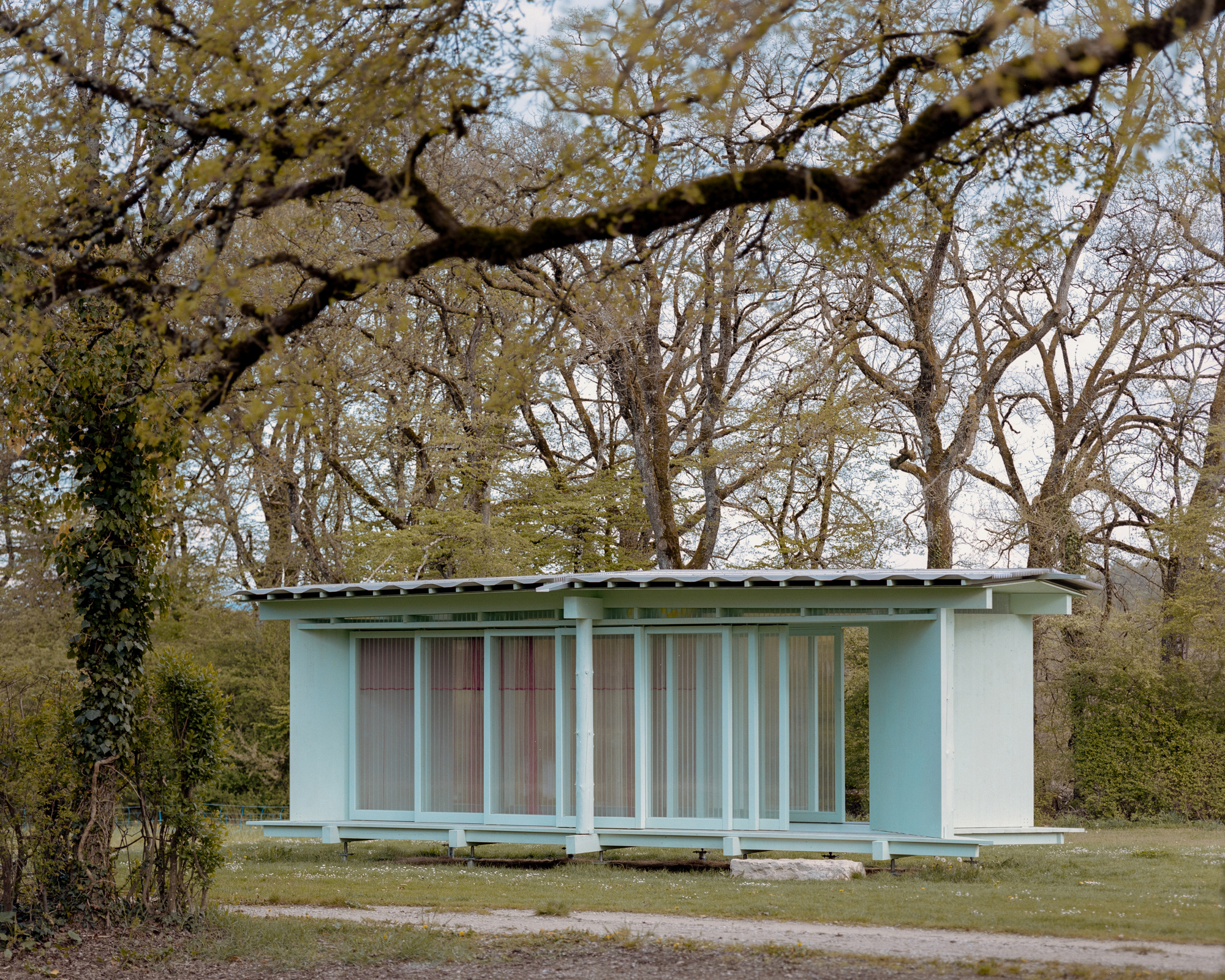 Side view of the pavilion through budding branches, with a gravel path crossing the foreground and ivy on a trunk