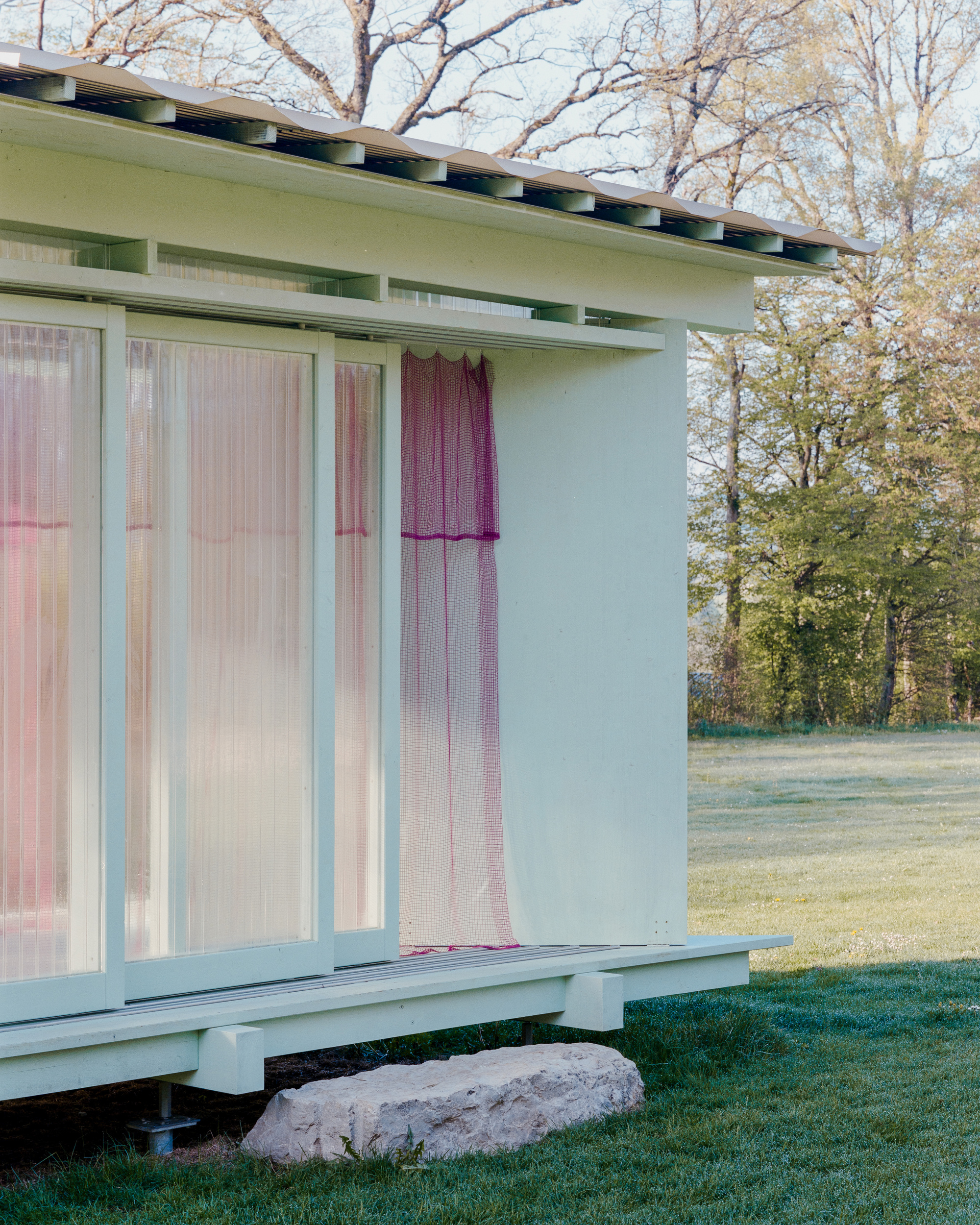 Close-up of the corner with exposed roof brackets, a draped magenta mesh curtain and a rough stone block on dewy grass
