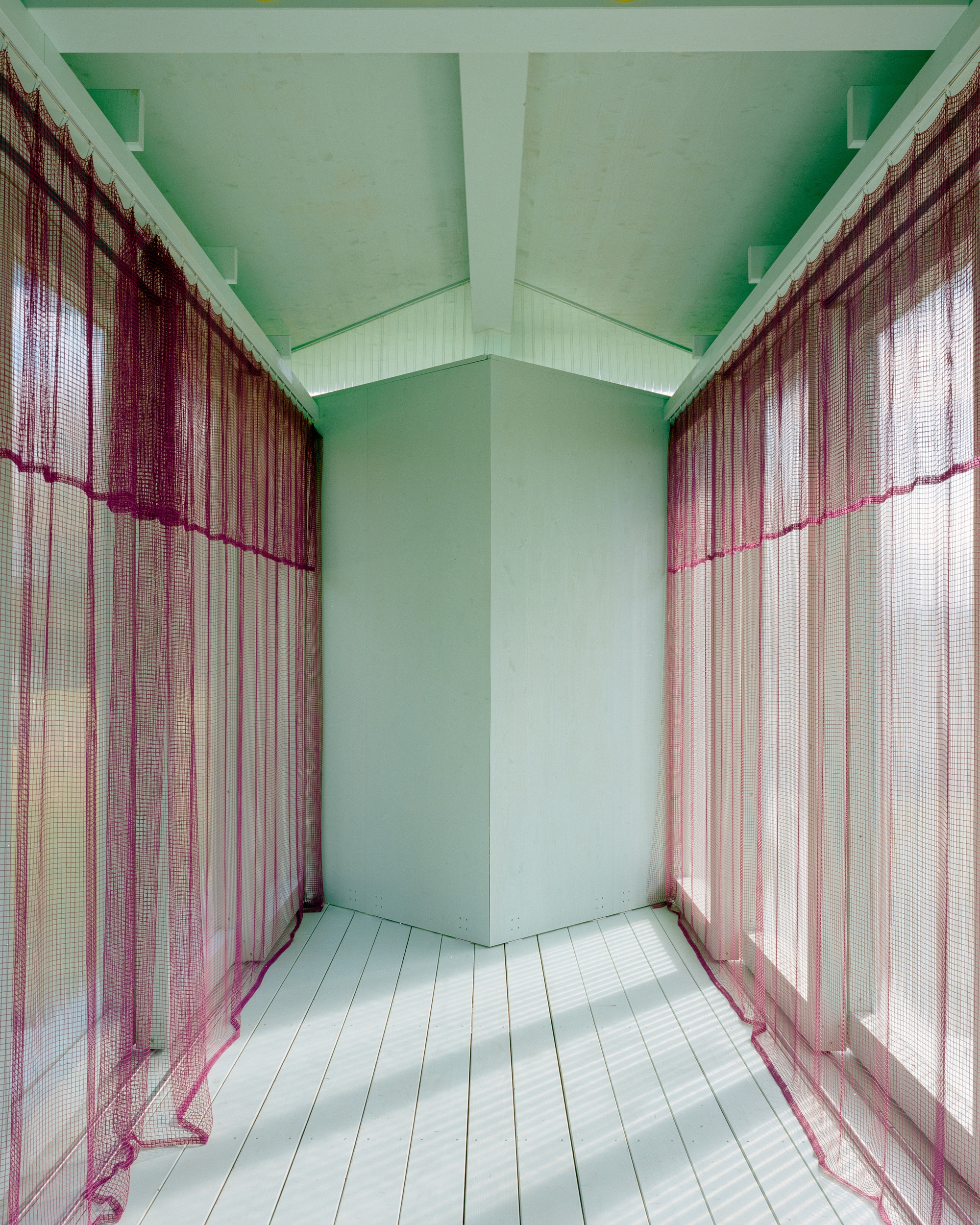 Narrow pale green room with magenta mesh curtains on both sides and sunlight falling across whitewashed floorboards